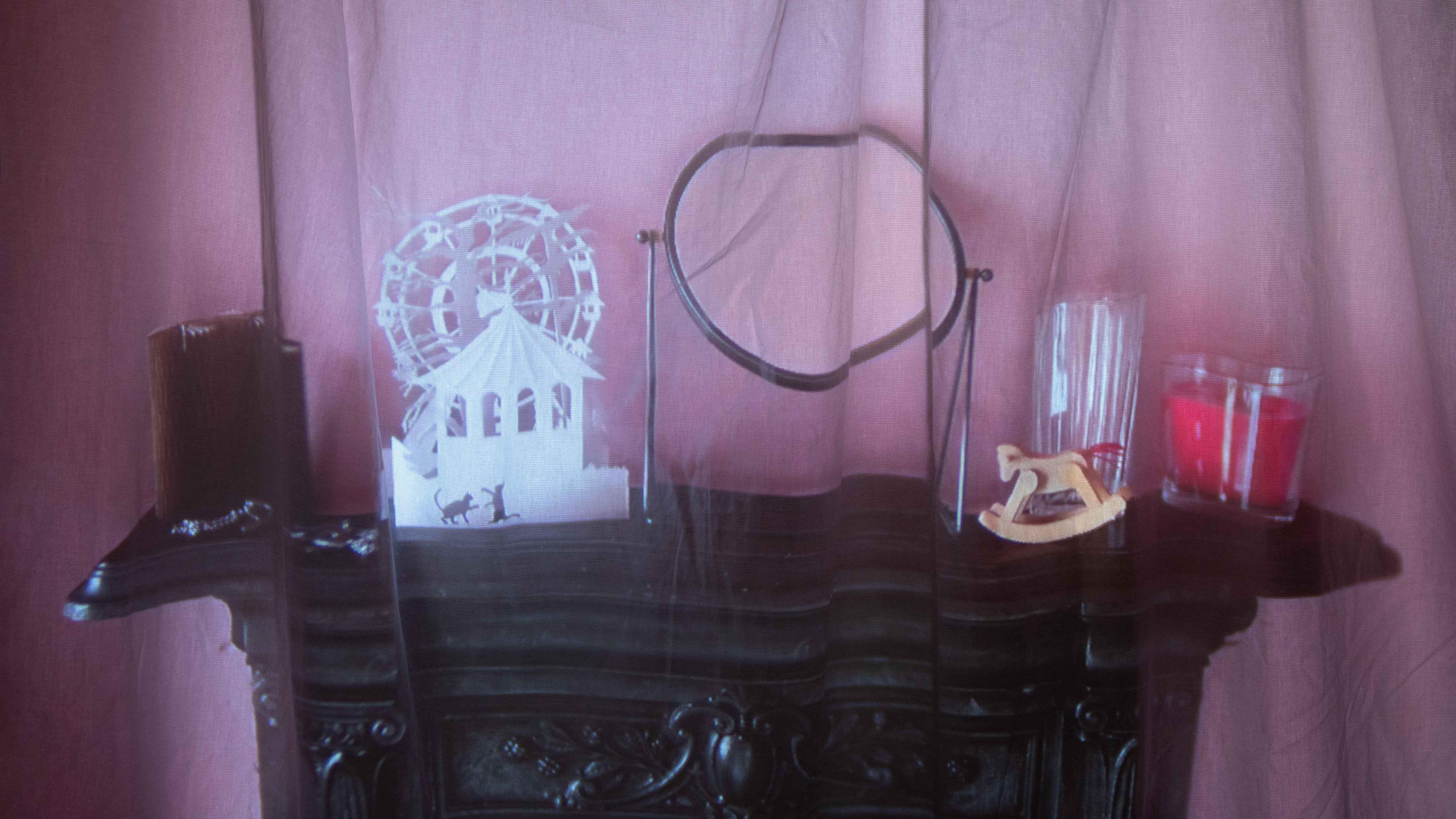 A black Edwardian cast iron fireplace in a bedroom, with a shelf, with items on it, including a house with a wheel behind, a mirror, a rocking horse and candles. The image is projected onto a cotton cloth. 