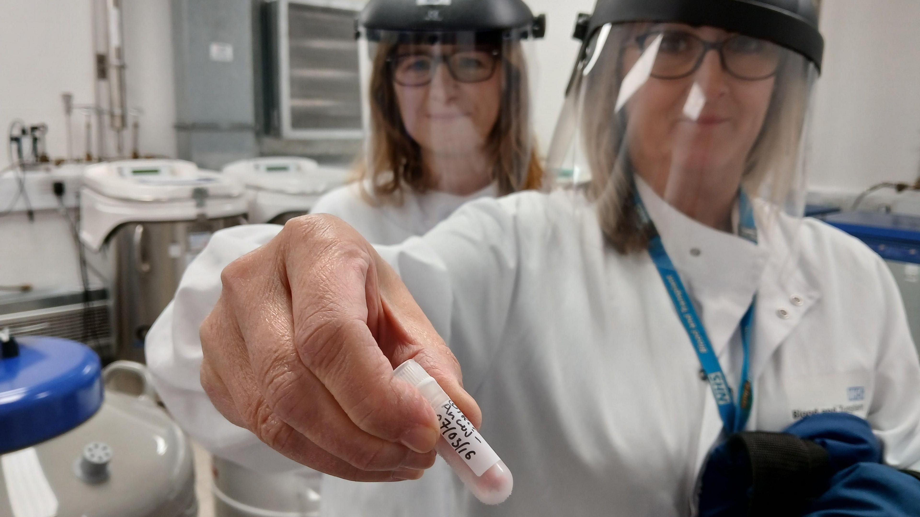 A woman wearing a white lab coat, holding a small capsule containing blood. A woman is stood behind her shoulder. Both are wearing PPE face guards.