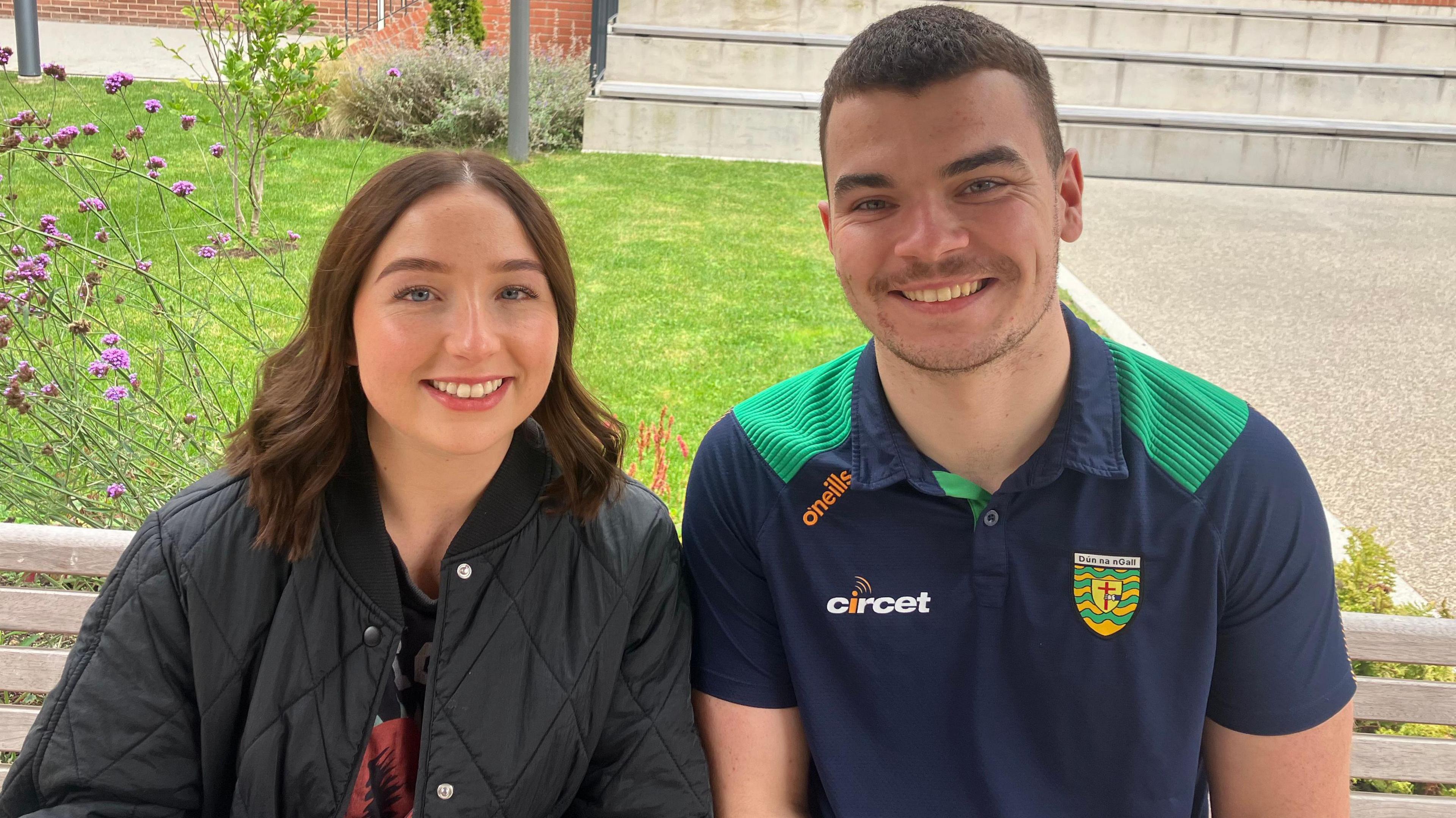 Eimhear O'Neill and Luke MacDaibheid sitting on a bench beside each other. They are both looking at the camera and smiling. Ms O'Neill is wearing a black coat and she has brown hair. Mr MacDaibheid is wearing a green and navy Donegal GAA polo. He has brown hair. 