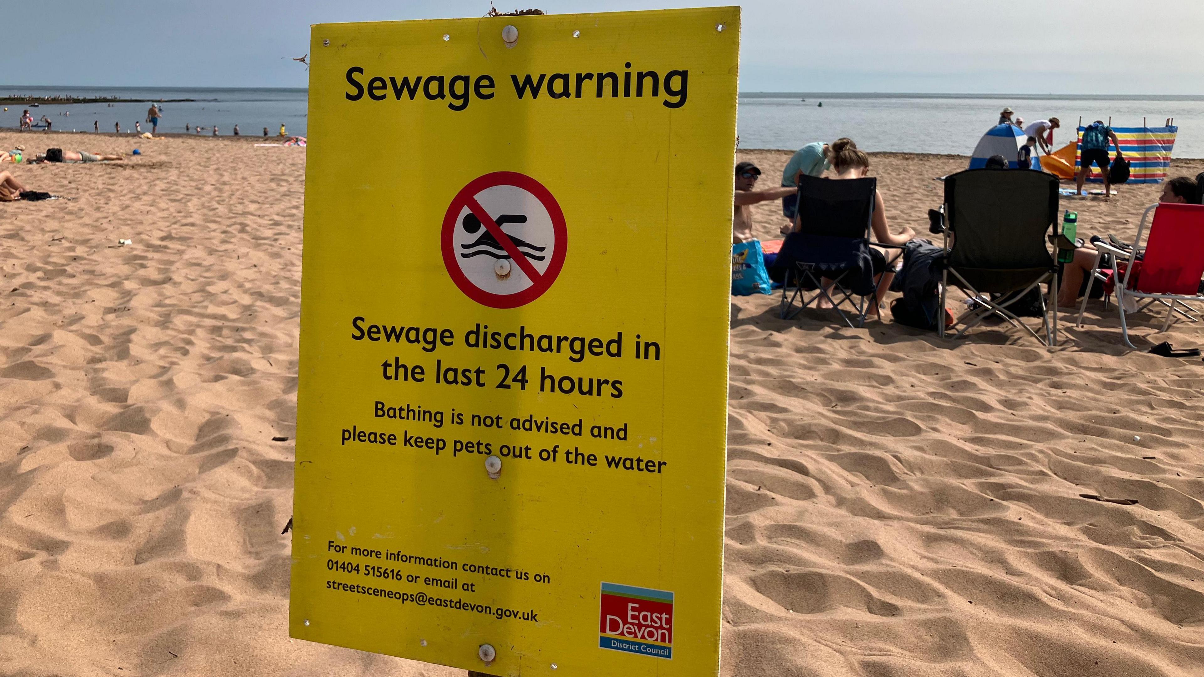 A beach scene with several people sitting on the sand facing the sea. In the foreground, a prominent yellow warning sign from East Devon District Council advises against bathing due to recent sewage discharge and warns to keep pets out of the water.