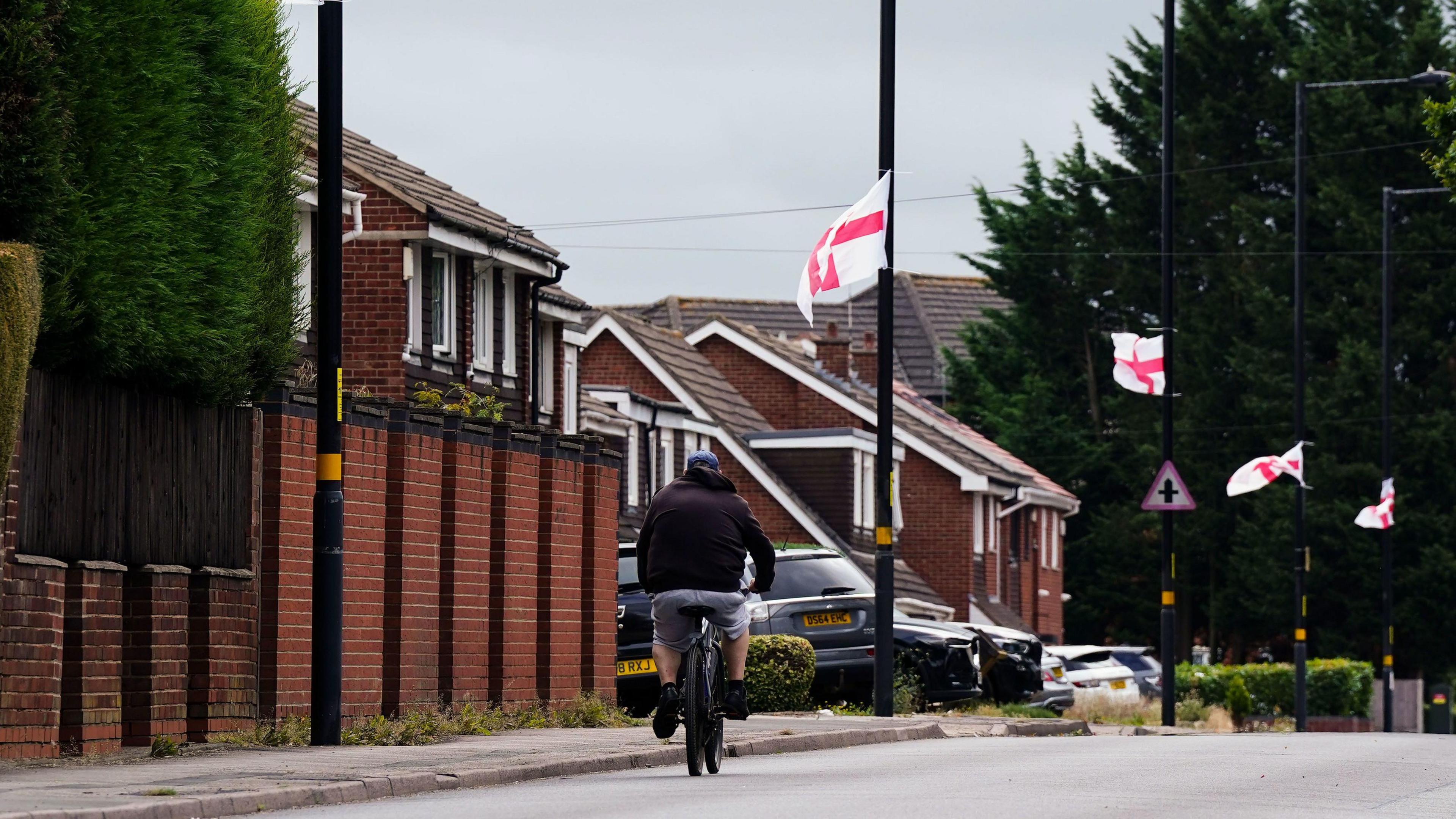 A person cycles down a residential street. Houses are on the left with cars parked outside. A number of St George's flags have been attached to several back lamp-posts along the pavement.