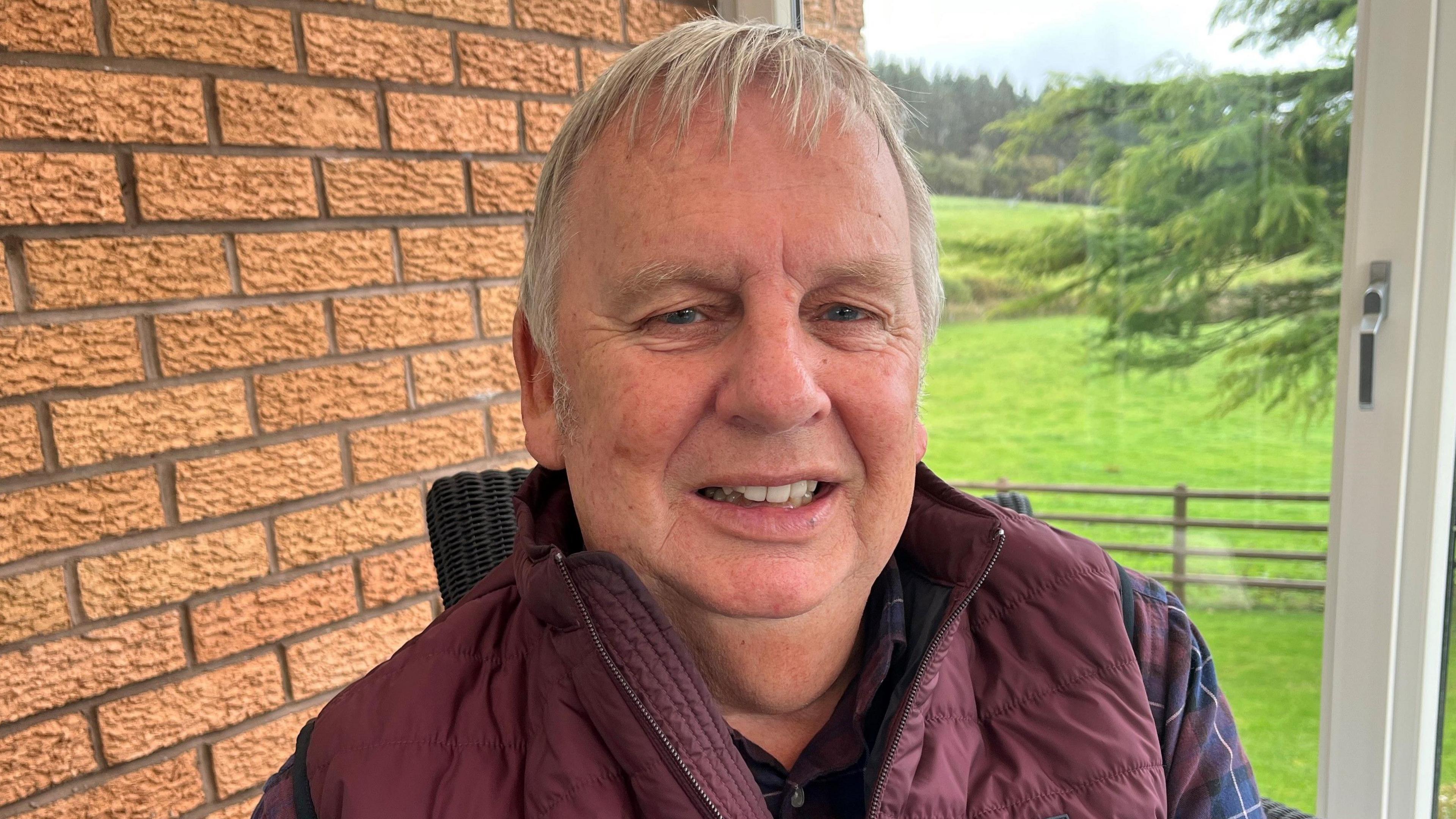 A head and shoulders shot of a man looking at the camera. He has short grey hair and a maroon gilet on over a blue and purple checked shirt. Behind him is a door with clear glass and outside you can see fields and the edge of a tree. There is a light brown bricked wall to the left behind the man. 