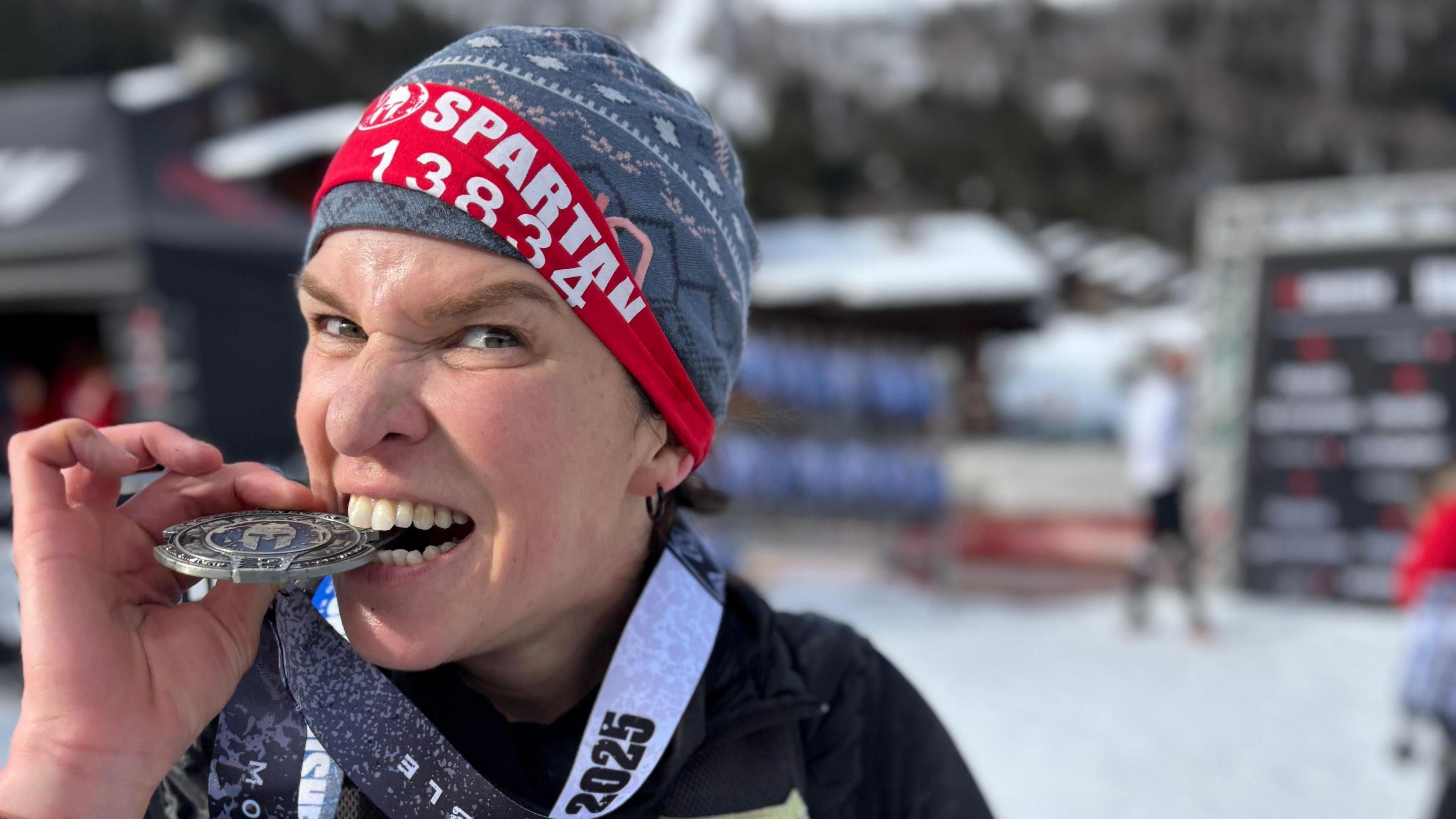 Fran Bullock biting her medal. She is wearing a hat and is looking at the camera. She has a red banner around the hat which has the word 'Spartan' on it with a number below.