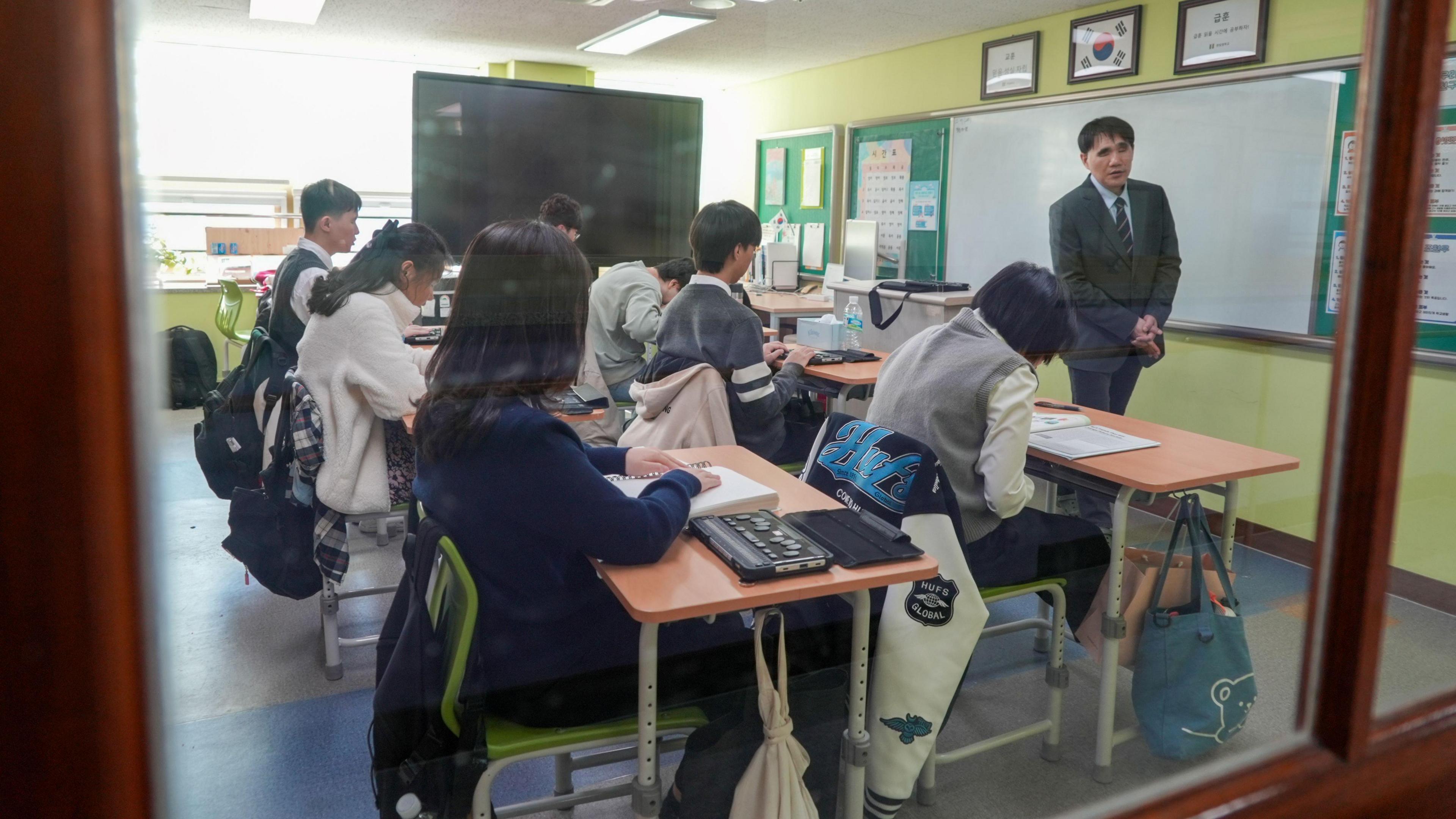 A classroom full of students at their desks, using braille materials and magnifiers, while a teacher stands in front of the whiteboard


