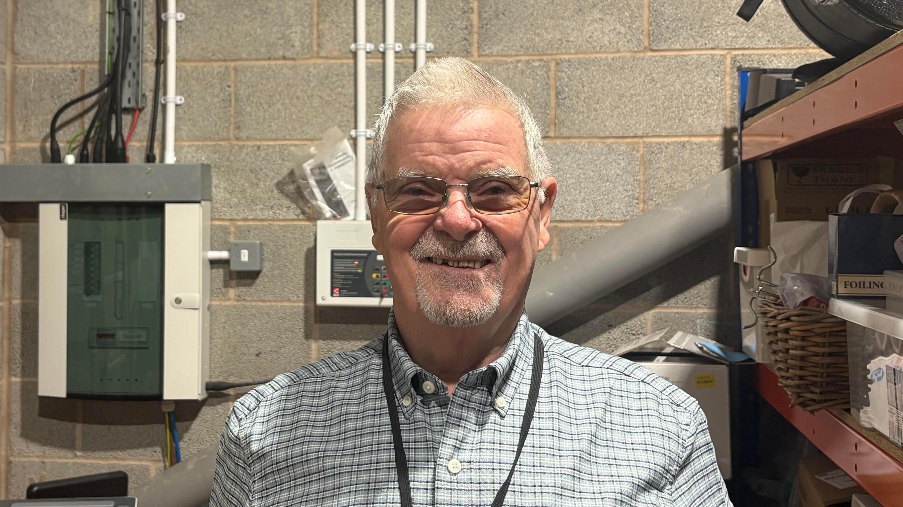 A head and shoulders photo of John Lydon smiling at the camera. He has short grey hair and a grey beard. He is wearing glasses, a blue checked shirt and a black lanyard.