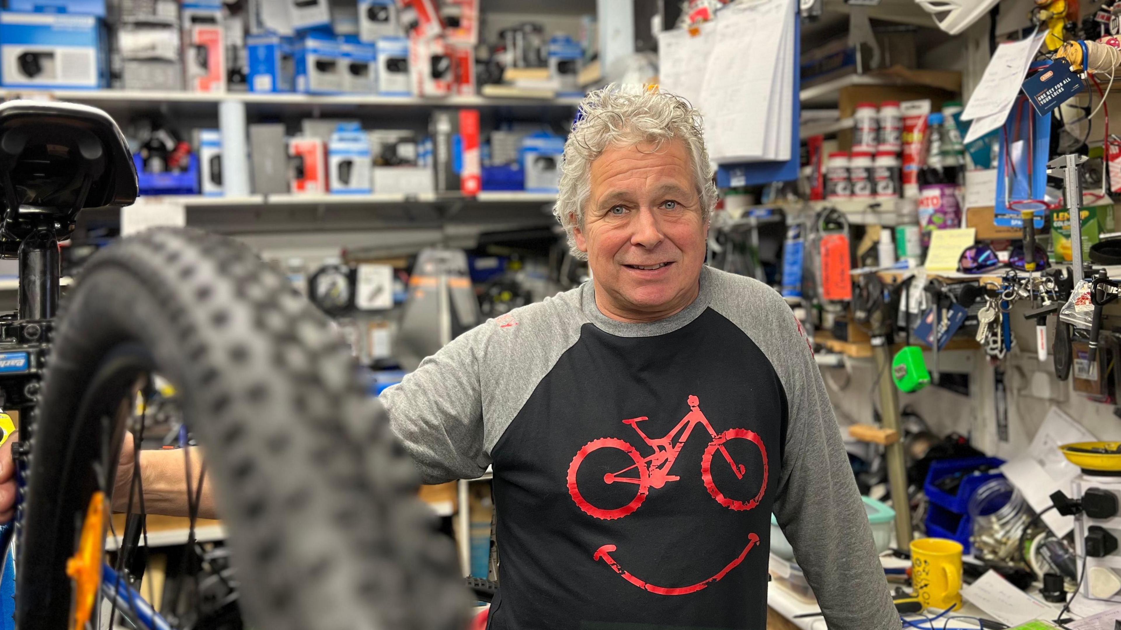 Aaron Lappage is standing in his bike shop. He's a man with curly grey hair. He is wearing a black and grey top with a red bike on it. He is smiling into the camera and leaning on a bike that is elevated, with its back wheel coming into the front of the photo.
