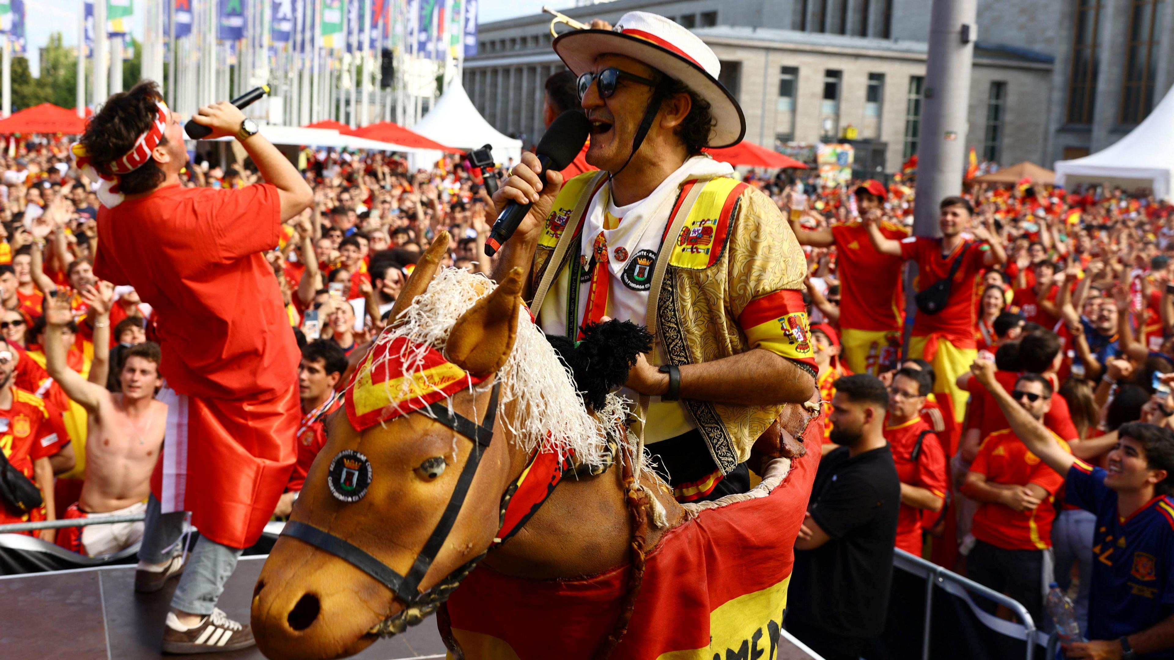 Man with a fake horse strapped to him speaks into a microphone on a stage in front of a crowd of Spain's fans wearing red and yellow