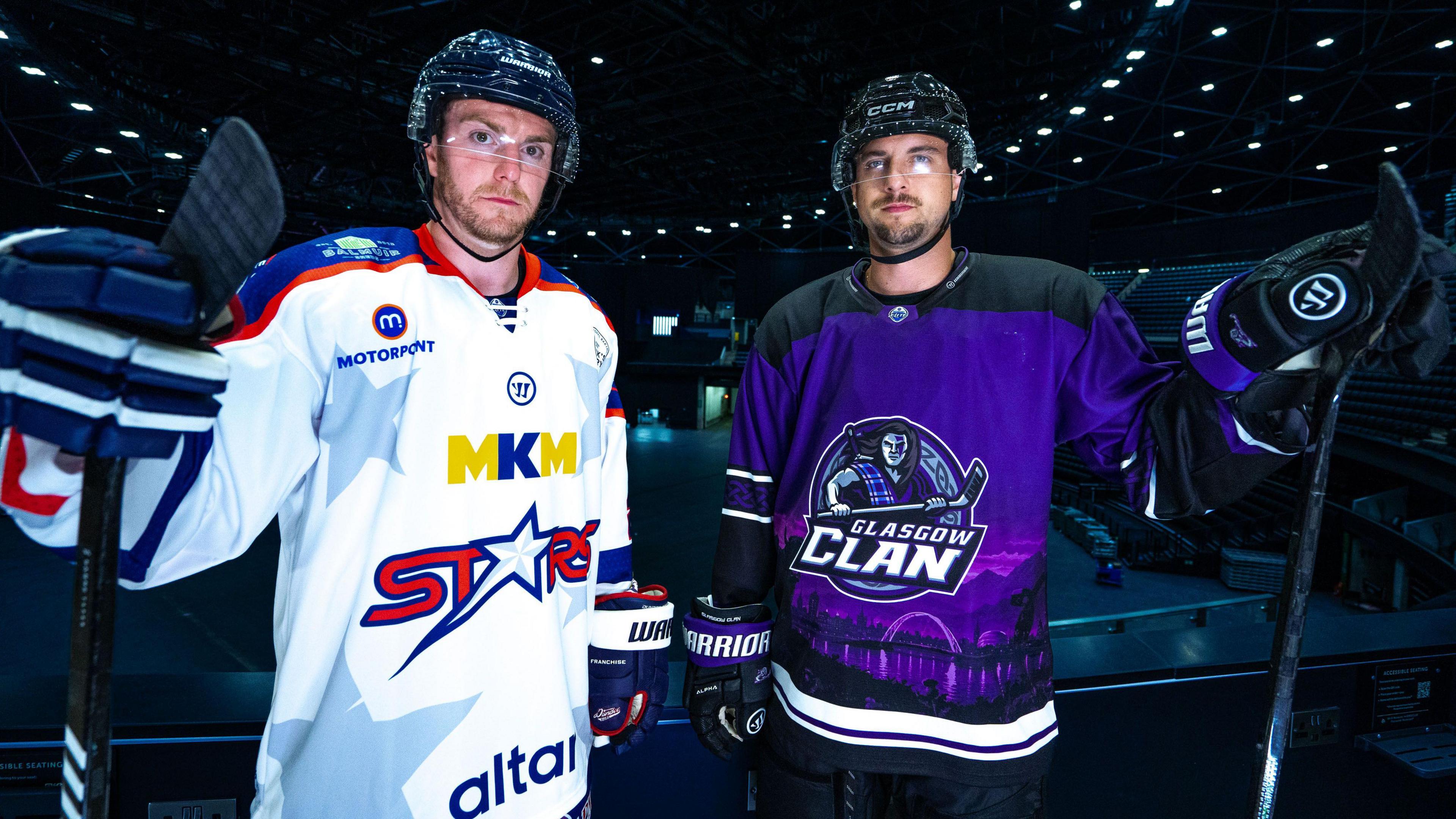 Two ice hockey players standing inside an arena. Craig Moore is wearing a white jersey with stars patterned on it, Deven Sideroff is wearing a purple and black jersey.