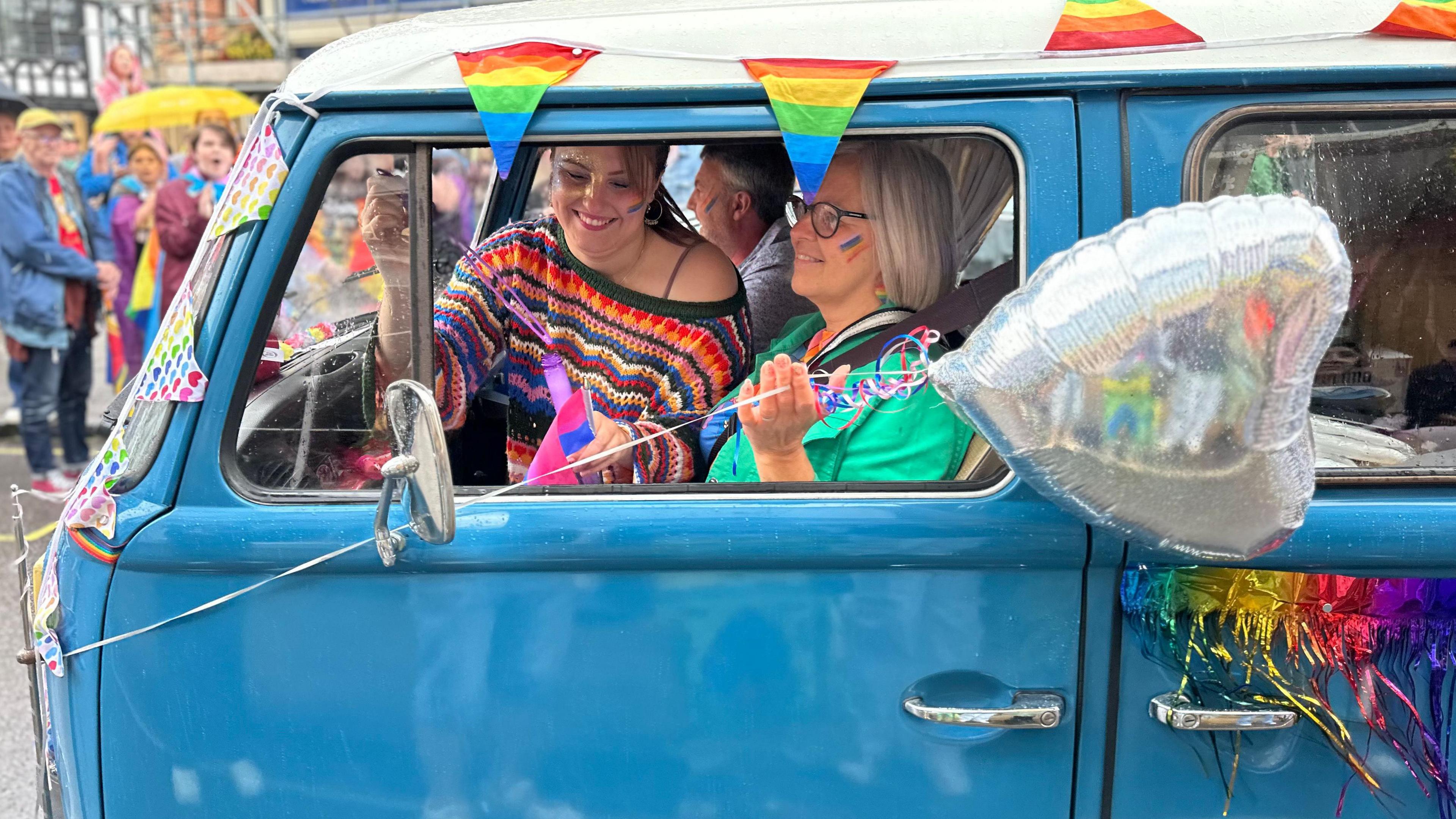 Smiling women in a vintage VW van, adorned with balloons and pride flags