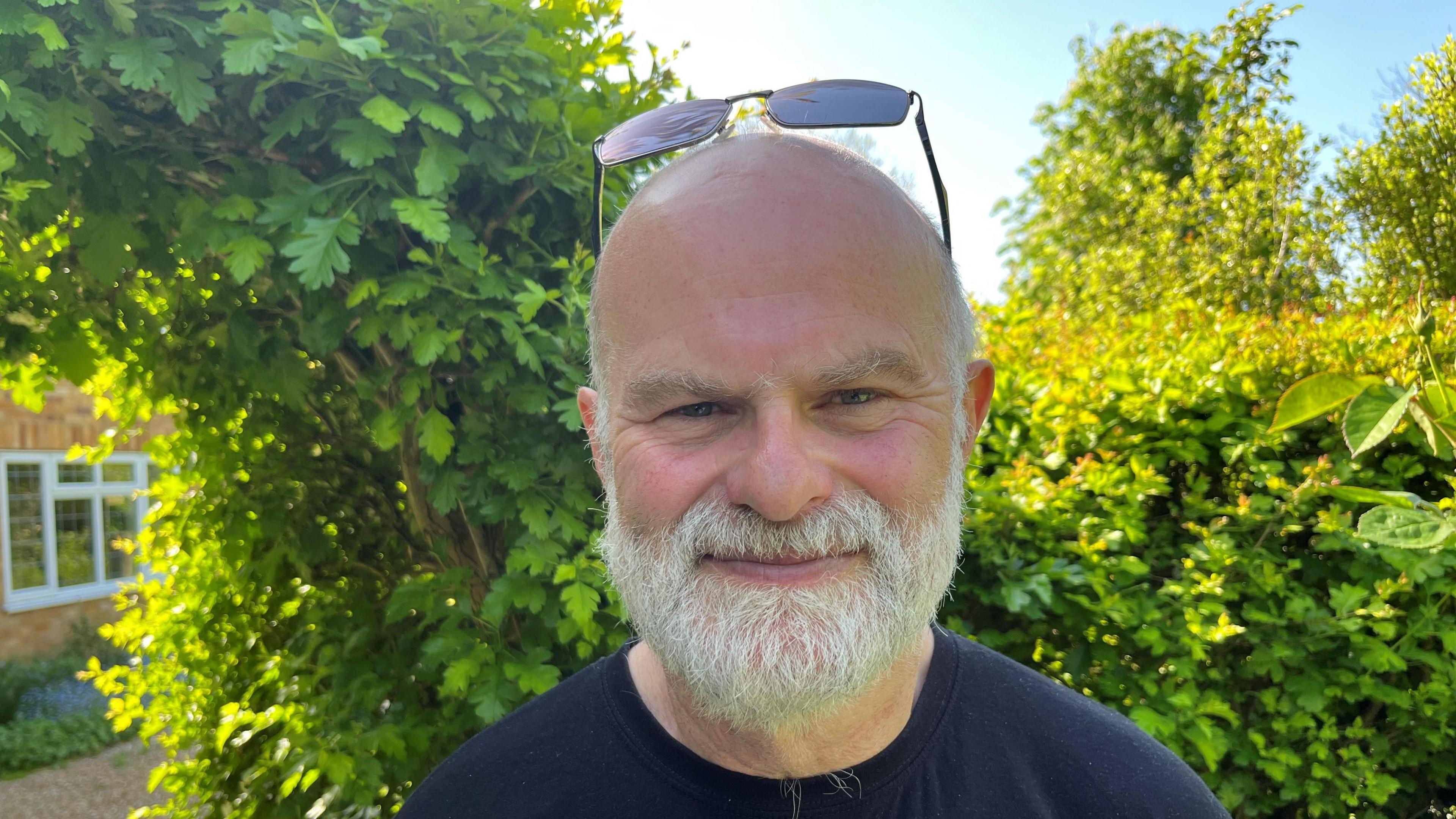 Andy Purvis, 62, has a bald head and a white/grey beard and moustache, with some hair on the sides of his head. He is wearing a blue jumper and has a garden hedge and house behind him.