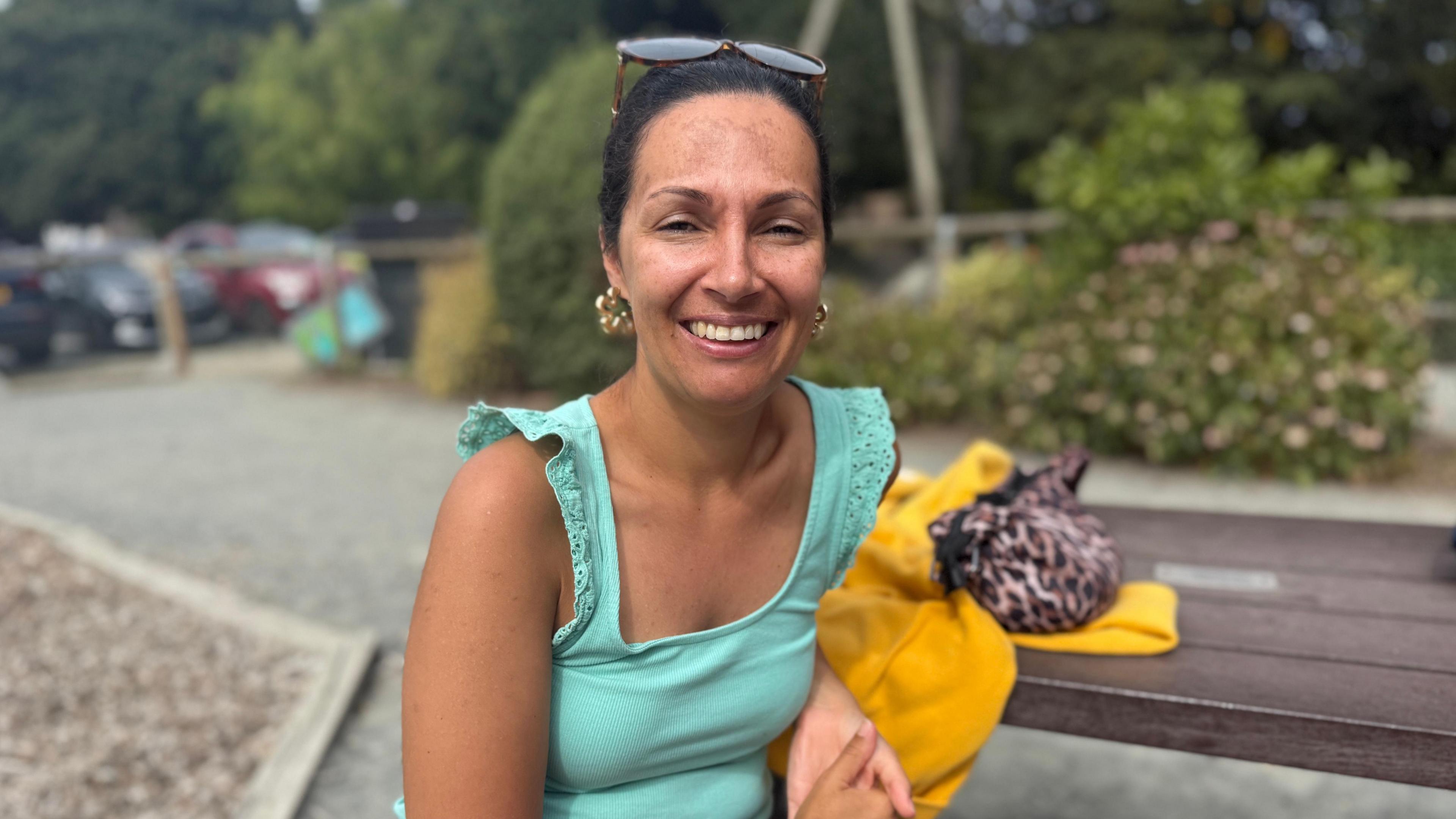 Women in her 30s in a blue top leant on a yellow jumper. She has brunette hair up in a claw clip and big gold earrings