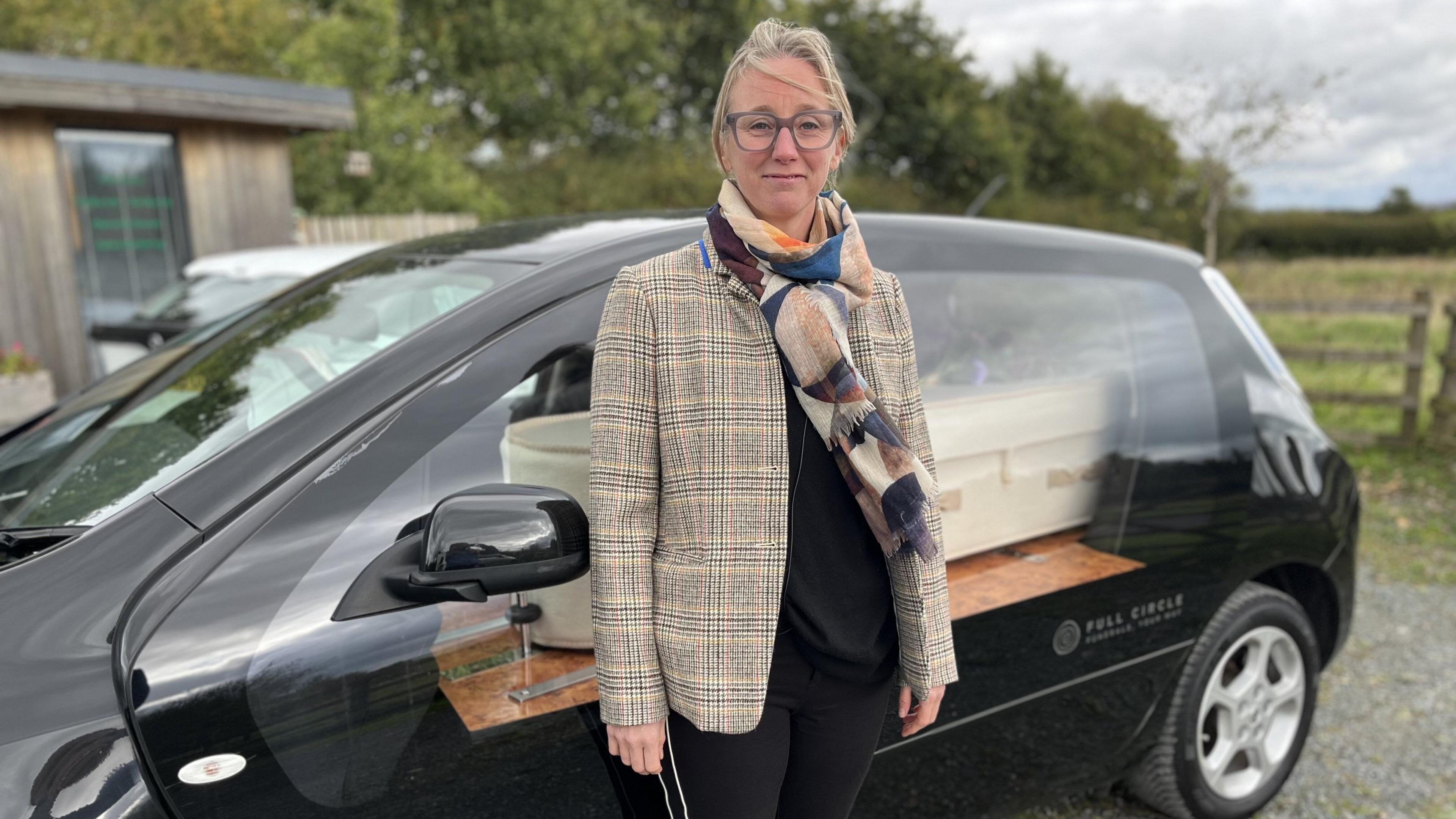 Sarah Jones has blonde hair tied back and is smiling at the camera. She wears glasses, a checked blazer, multicoloured scarf and black t-shirt and trousers. She is standing in front of a black electric hearse which has a big glass panel instead of separate windows. Behind the glass is a white wool coffin.