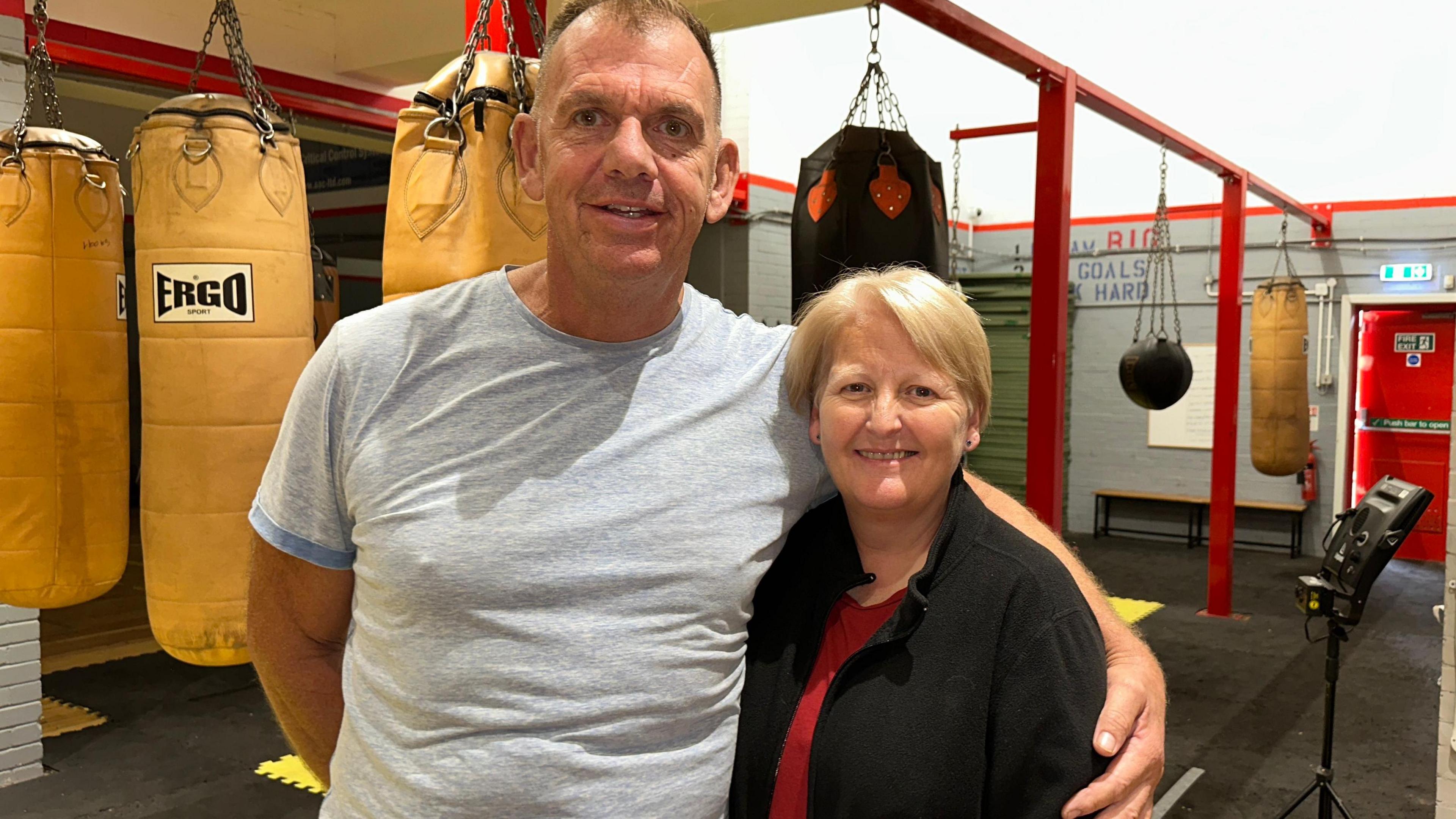 Man with short brown hair wearing a grey t-shirt and woman with short blonde hair wearing a black cardigan and red top and both smiling, standing in a boxing gym with punchbags in the background. 