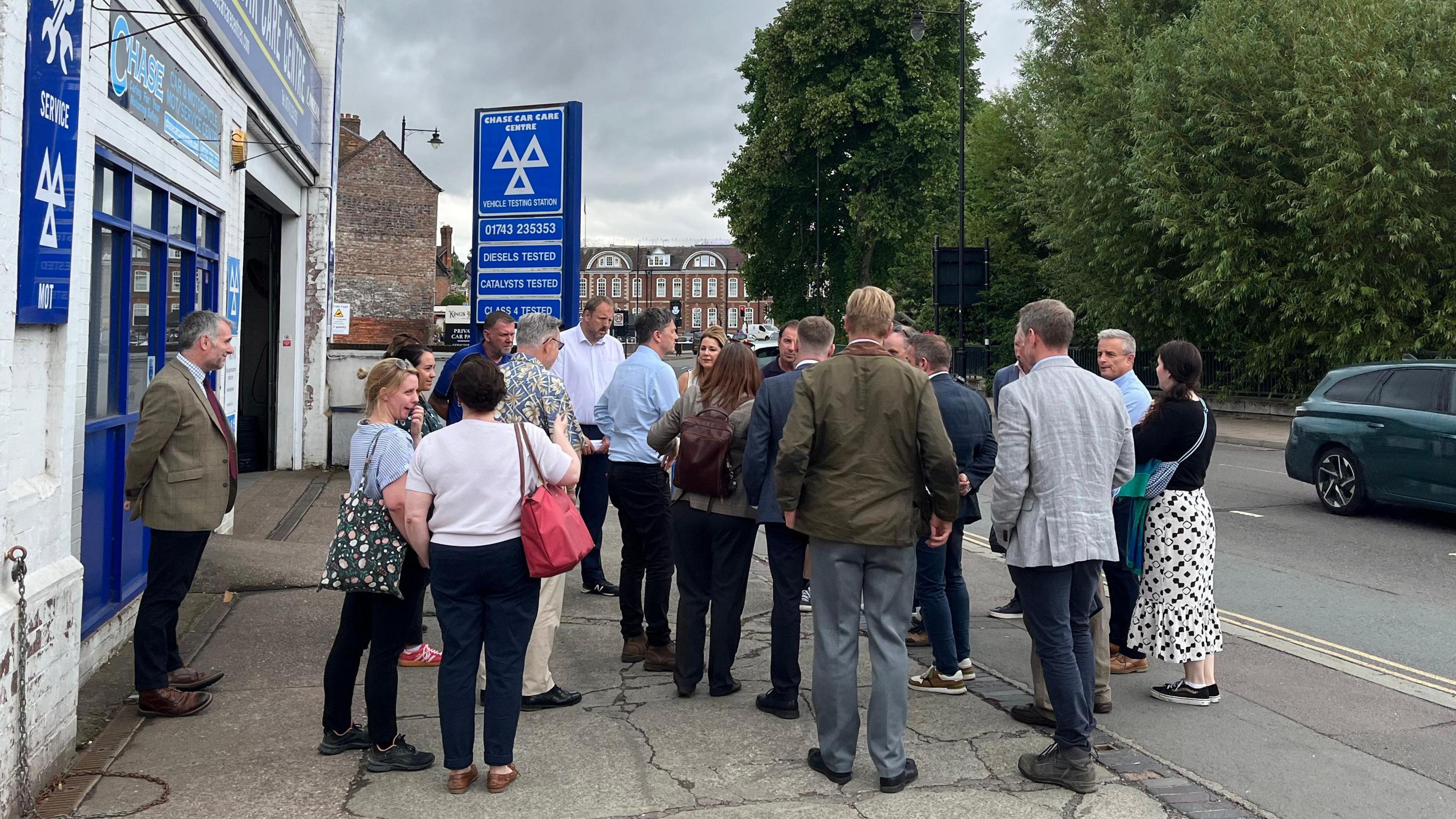A group of around 20 people stood on the forecourt of Mark's business, a single-storey whitewashed building with a blue sign above it - in white letters the company name is written - Chase Car Care Centre. The group are facing away from the camera and talking to one another.