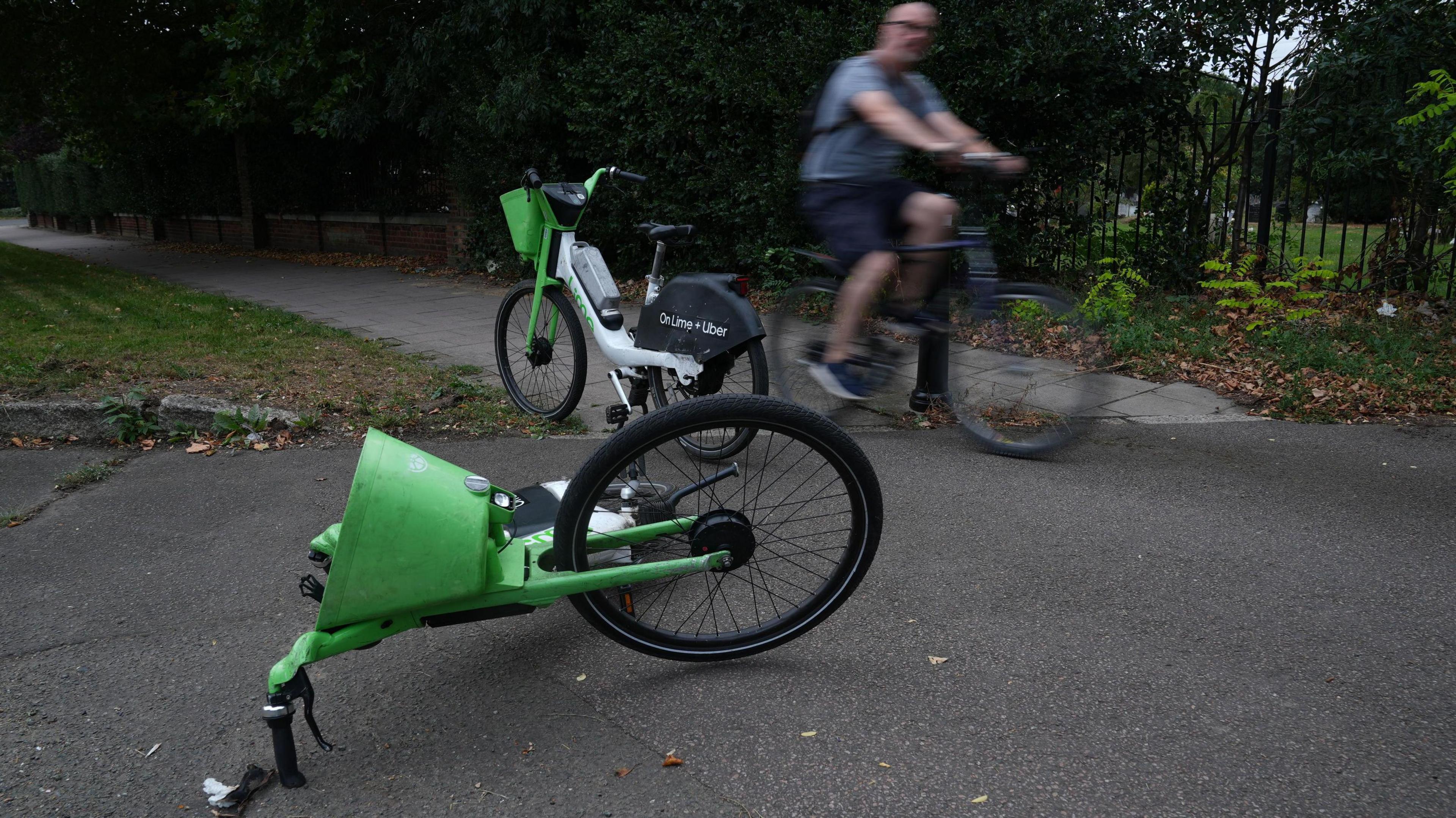 Two Lime bikes on a path, one standing and the other on its side, by a park. A cyclist is on his bike passing by in the background.