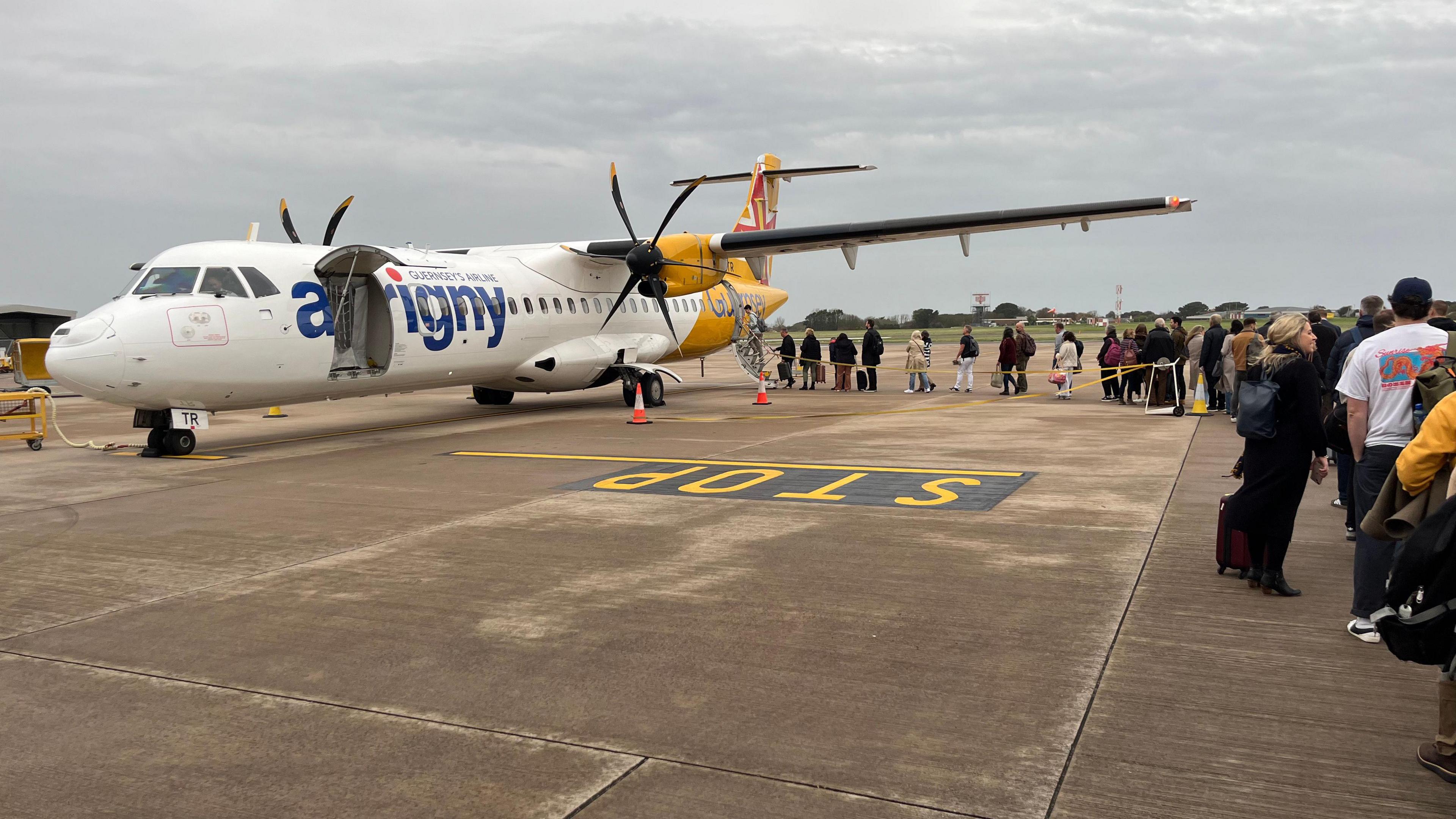 Dozens of people line up on the tarmac to board an Aurigny plane. The plane has yellow and white livery. It has two propellers. The passengers are entering through a door at the rear of the aircraft.