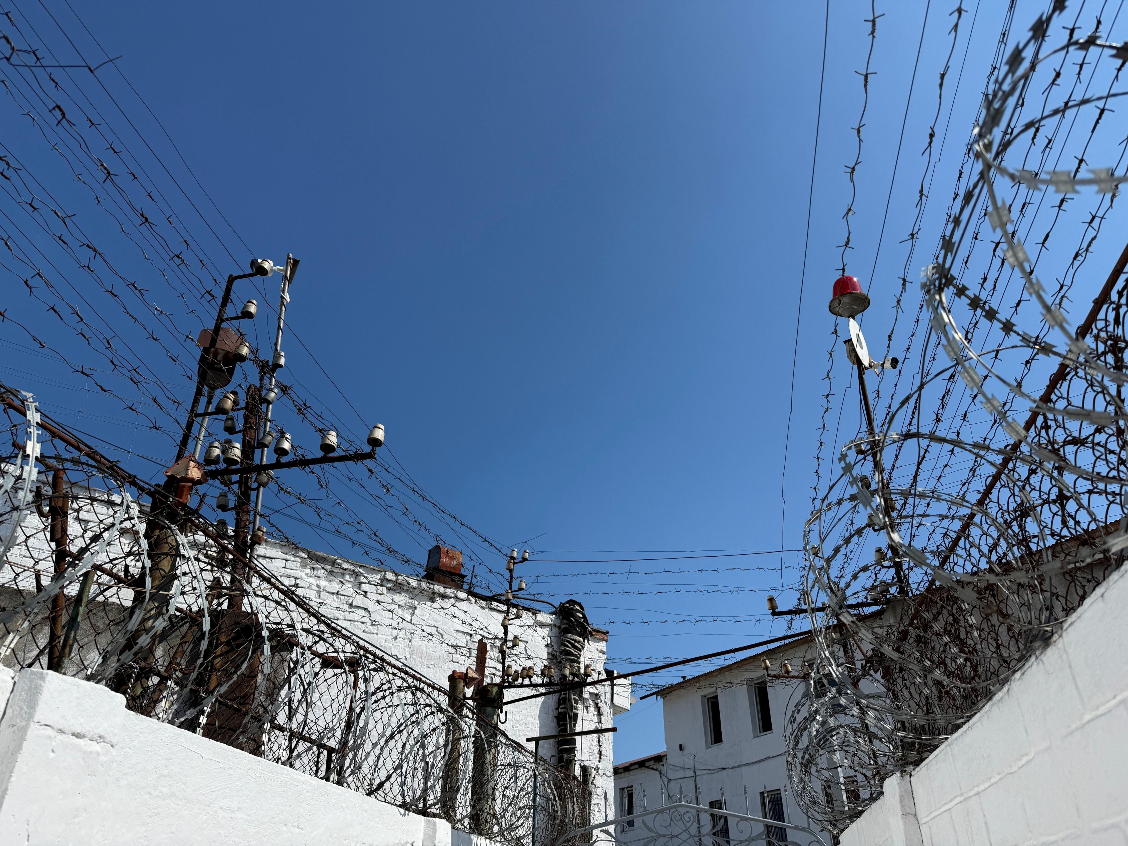Blue sky above white walls covered in barbed and razor wire. 