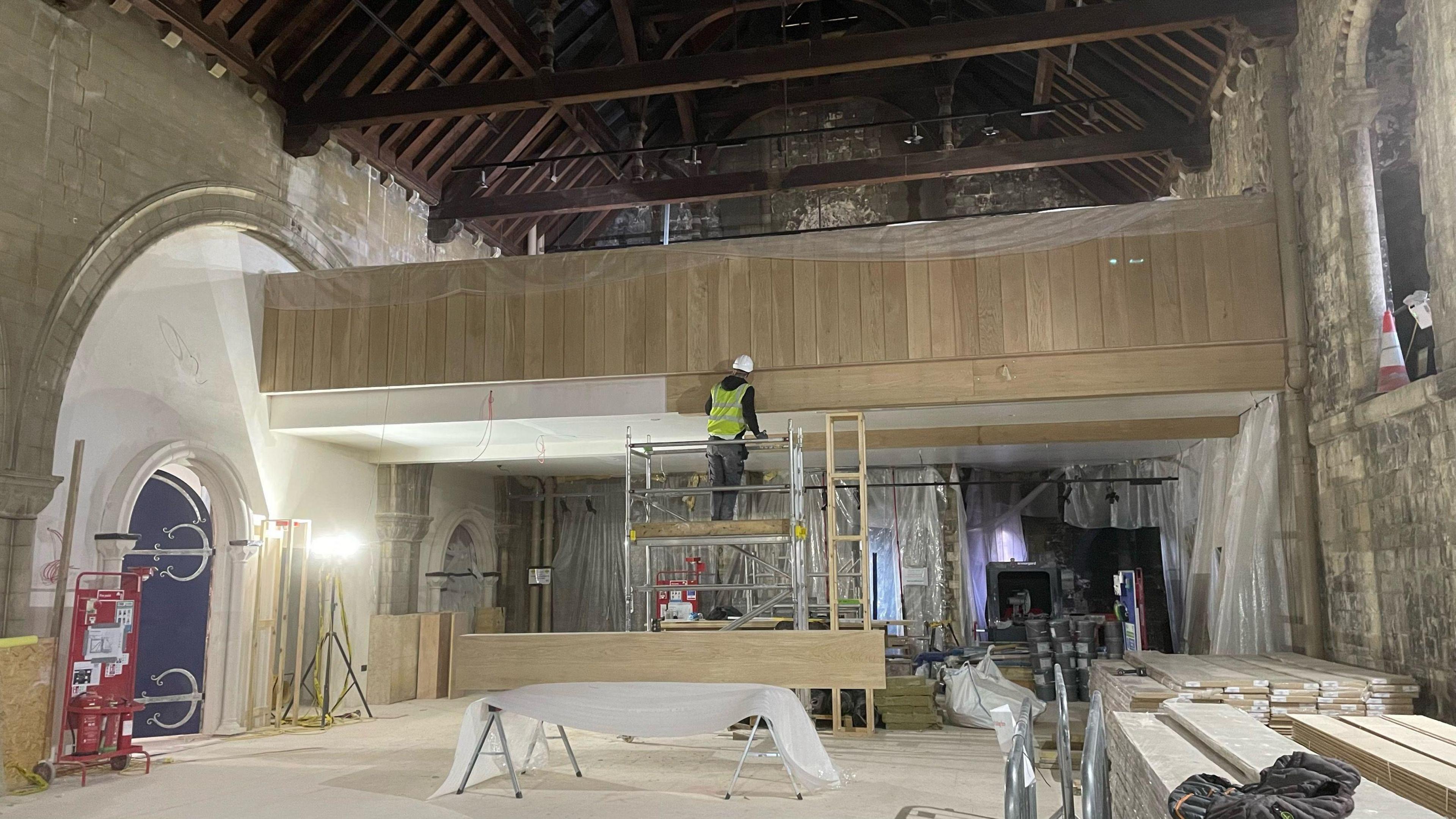 A workman stands on a scaffolding platform as he continues work inside Norwich Castle's keep. He is working on what appears to be a section of wooden panelling. The vaulted ceiling of the keep is above him. 