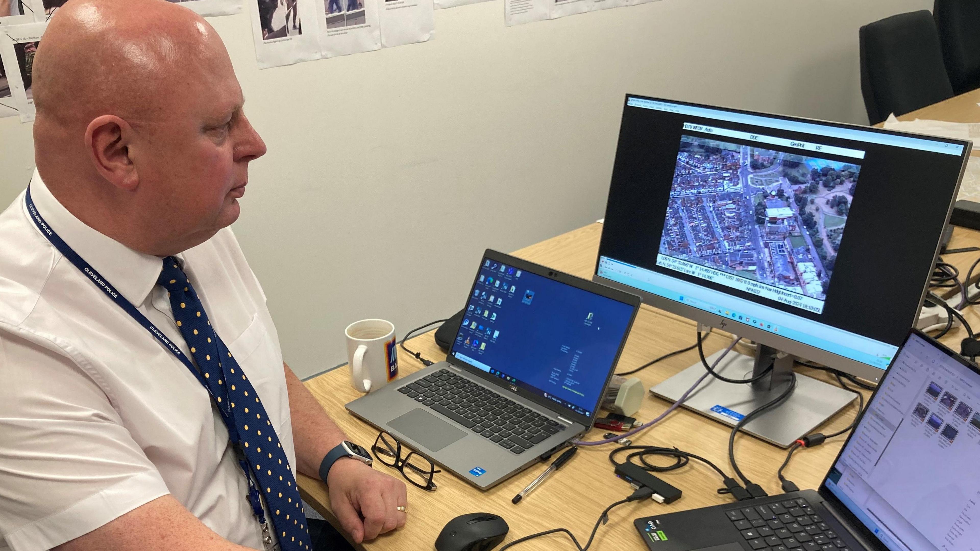 A side view of a man wearing a short sleeved shirt and tie seated at a desk with two laptops and a computer screen showing an aerial view of streets.