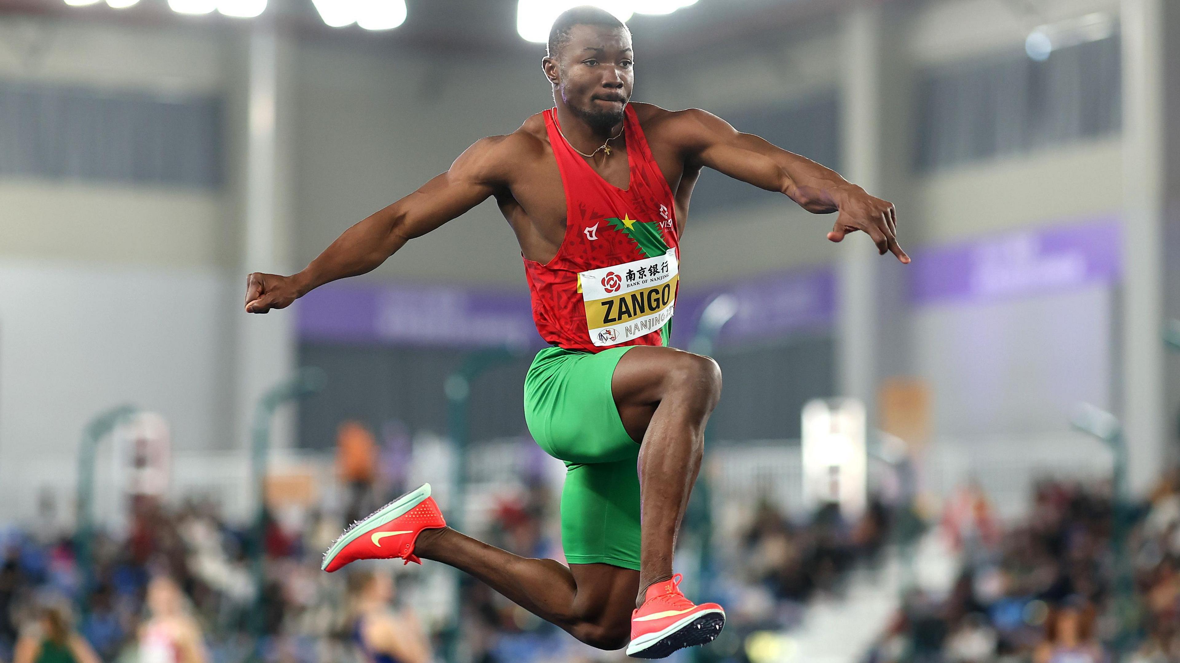 Hugues Fabrice Zango, wearing a red athletics vest, green spandex shorts and orange spiked shoes, is seen leaping in mid air during a triple jump event. A crowd is seen out of focus in the background