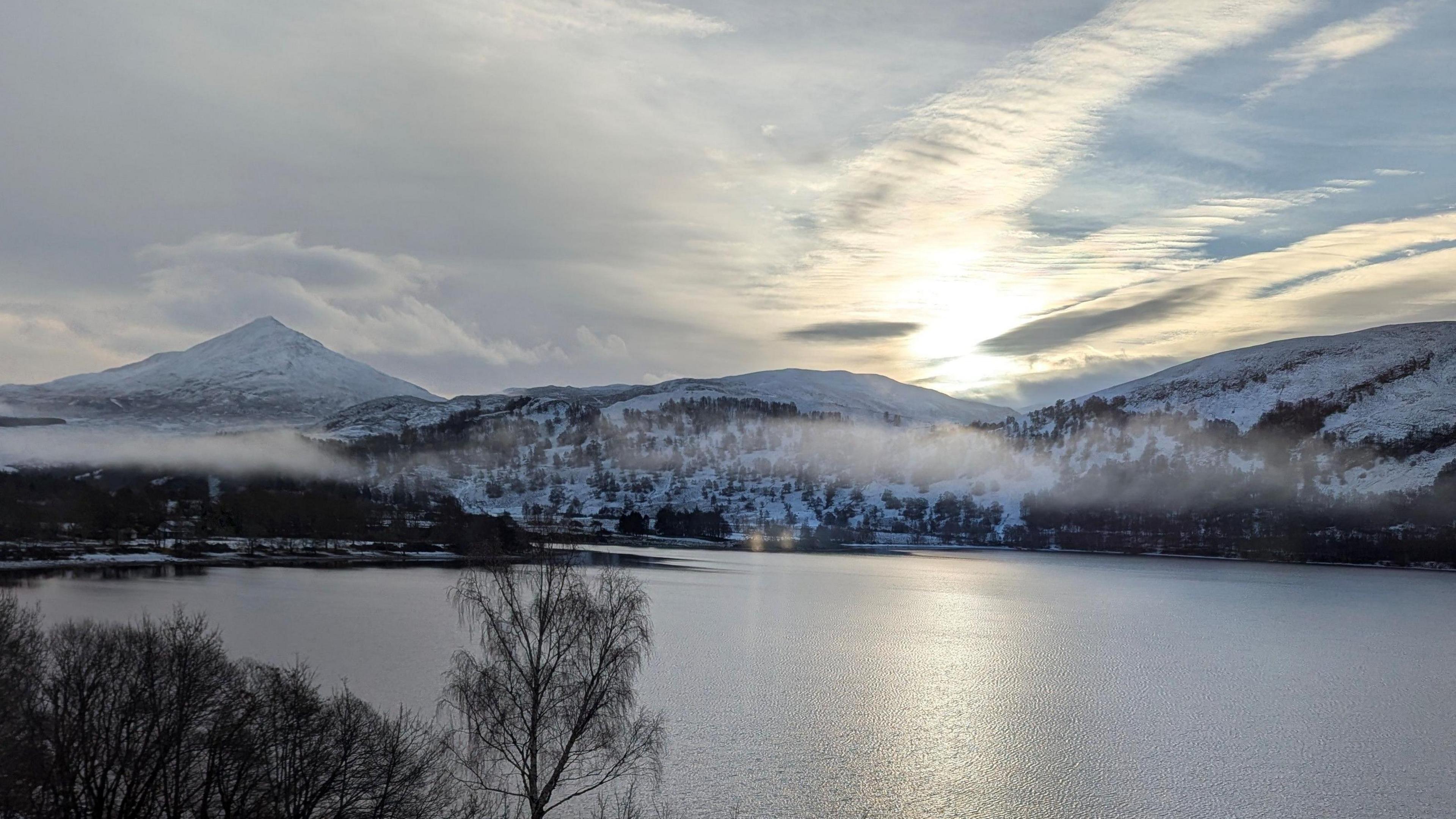 A view of Schiehallion from the window of a hotel room. The mountains are covered in snow. There are trees on the landscape which look dark. The sun is coming up.