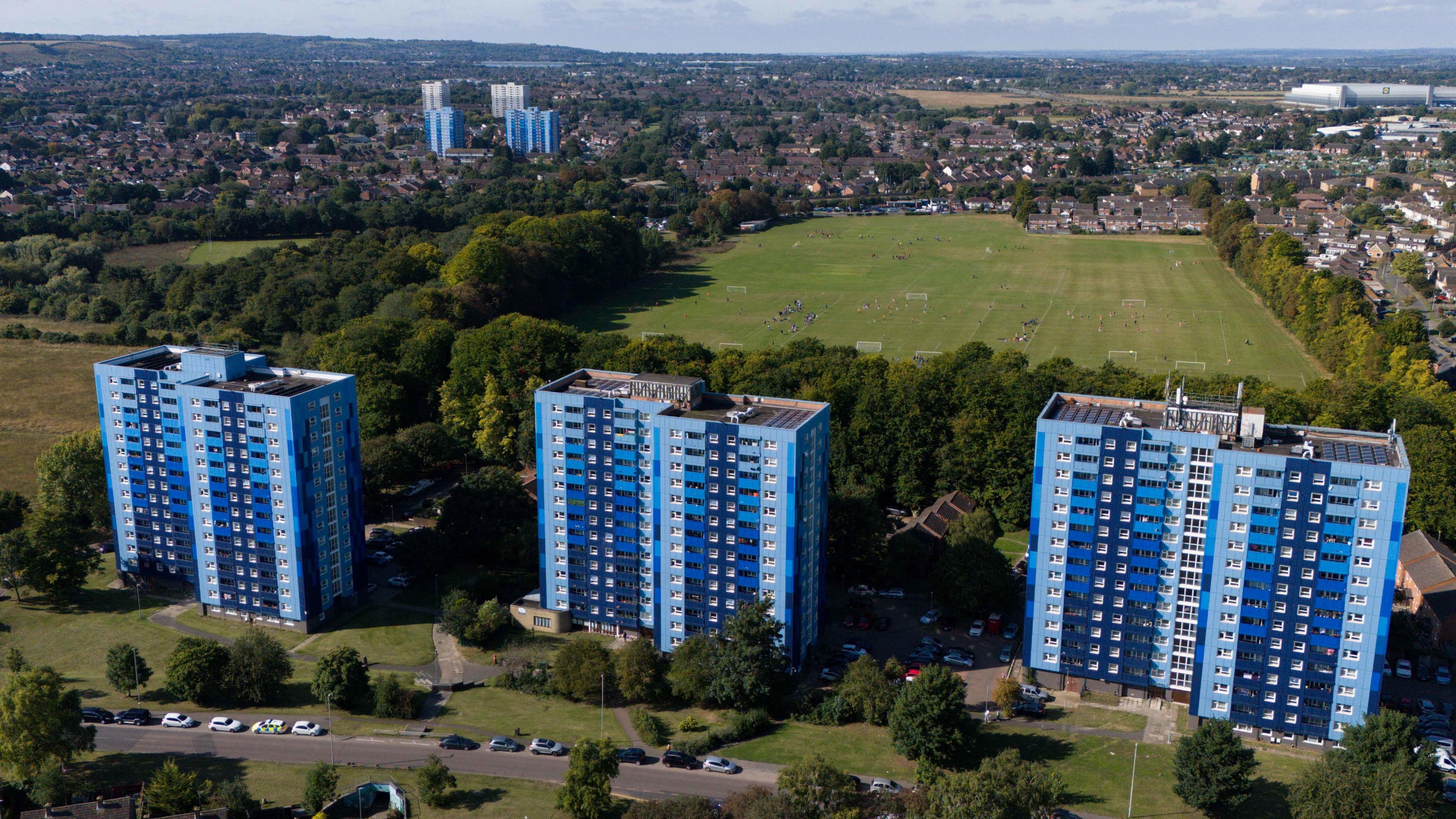 Aerial shot of three tower blocks, all in different shades of blue. Behind them is a large wooded area and an open expanse of playing fields, and more housing and tower blocks.