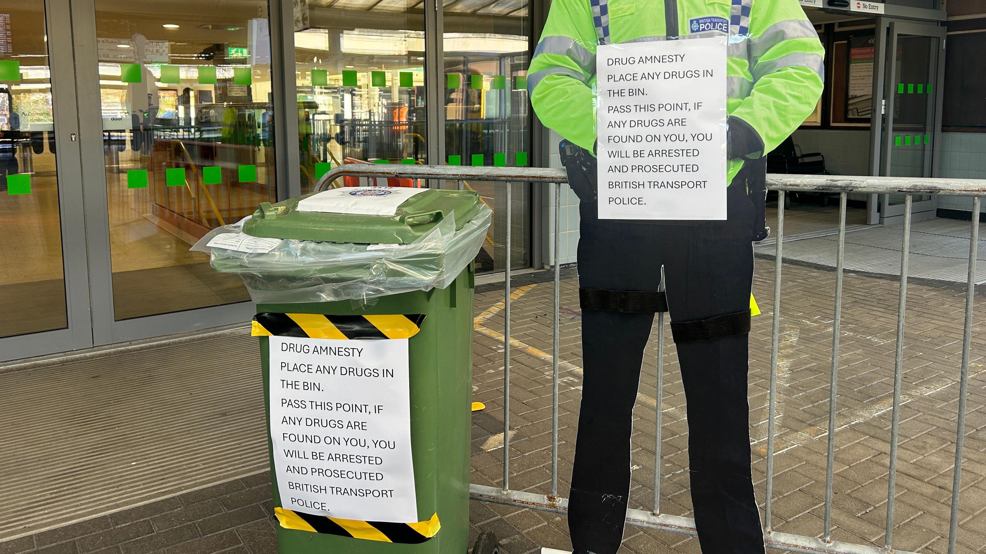 A green drug amnesty bin. A sign displayed on the front of the bin says drug amnesty place any drugs in the bin. Pass this point, if any drugs are found on you, you will be arrested and prosecuted British Transport Police. A life size cardboard cut out of a police officer is holding a sign with the same warning.