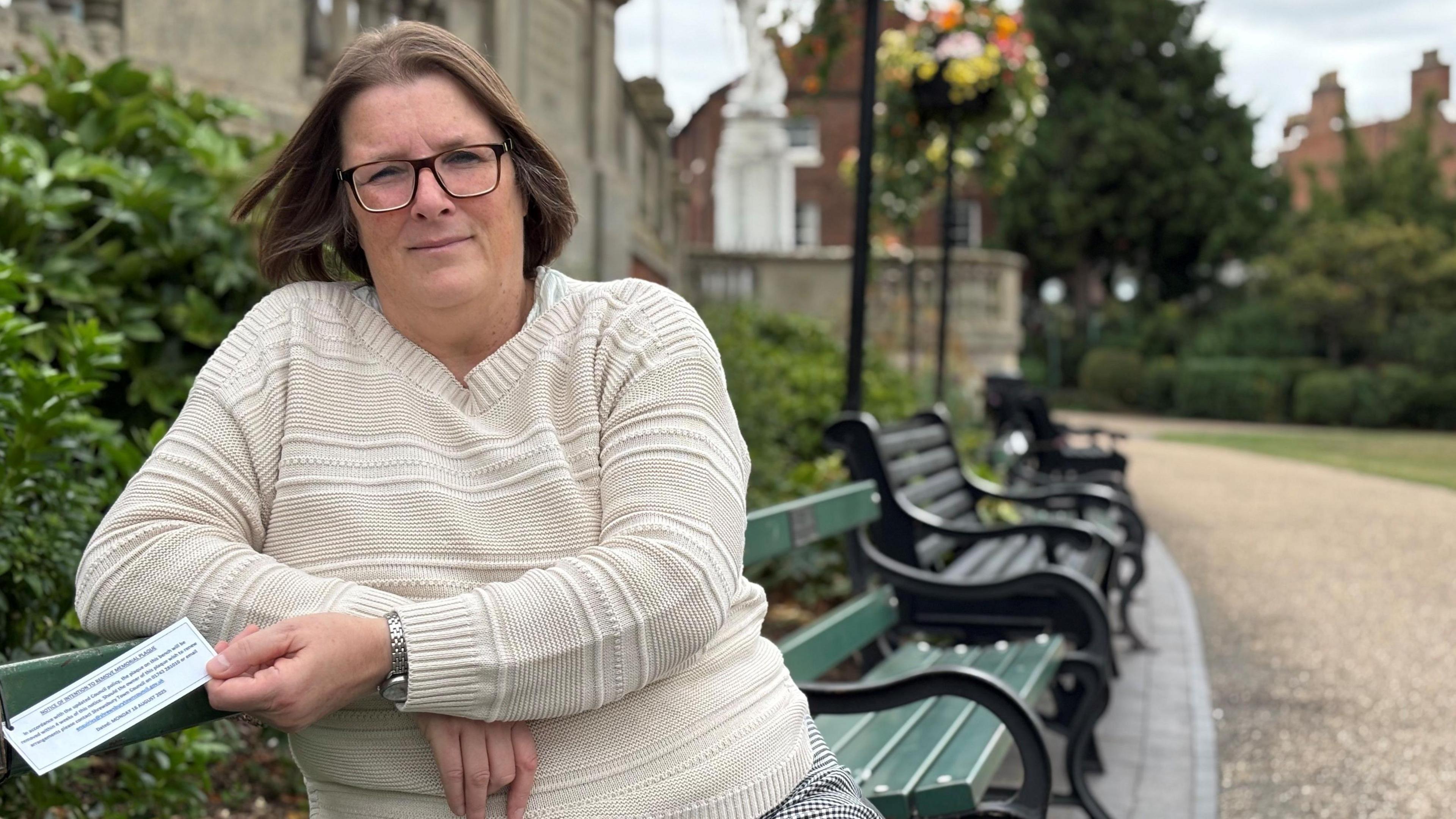 A woman wearing a white woollen jumper is sitting on a green park bench. She's holding a white sign, which is tied to the bench. There are buildings, a path and hanging baskets in the background.  