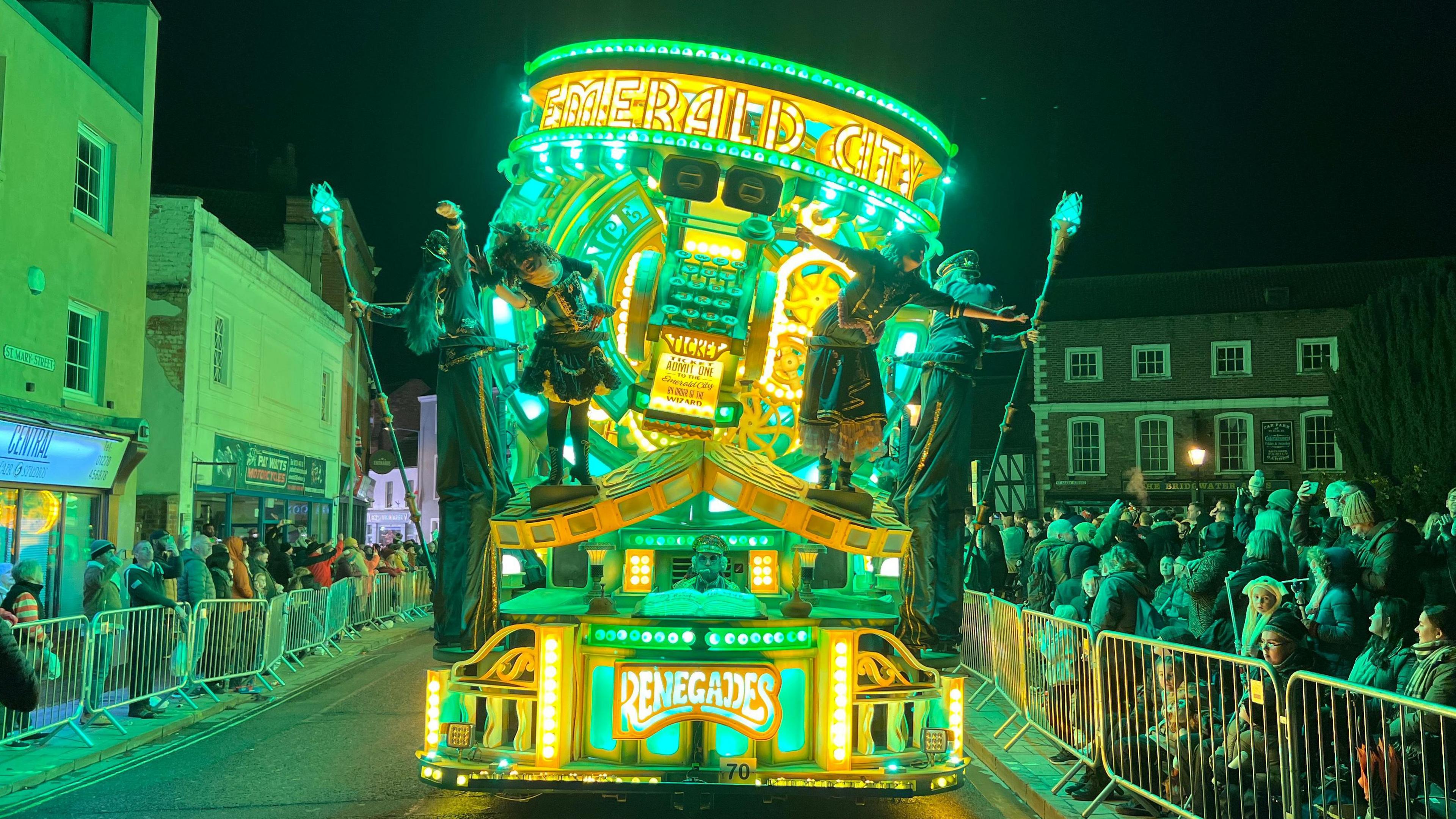 Big cart which is full of green and yellow LED lights shining. There's performers hanging off it. It's got the Renegades logo at the bottom. The cart is going through the street with people lining the street watching it.