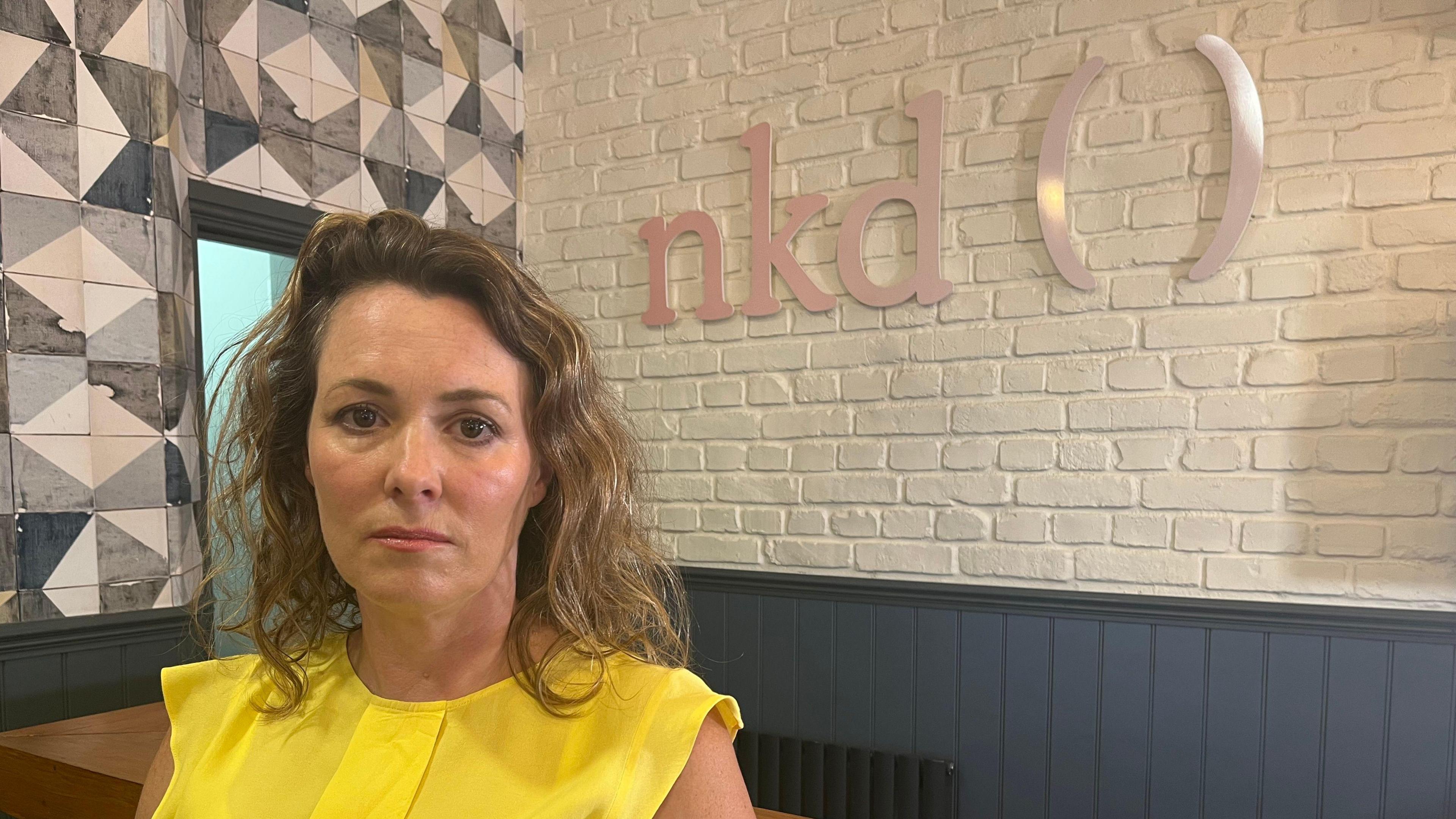 Woman wearing a bright yellow top standing in front of a sign which read nkd