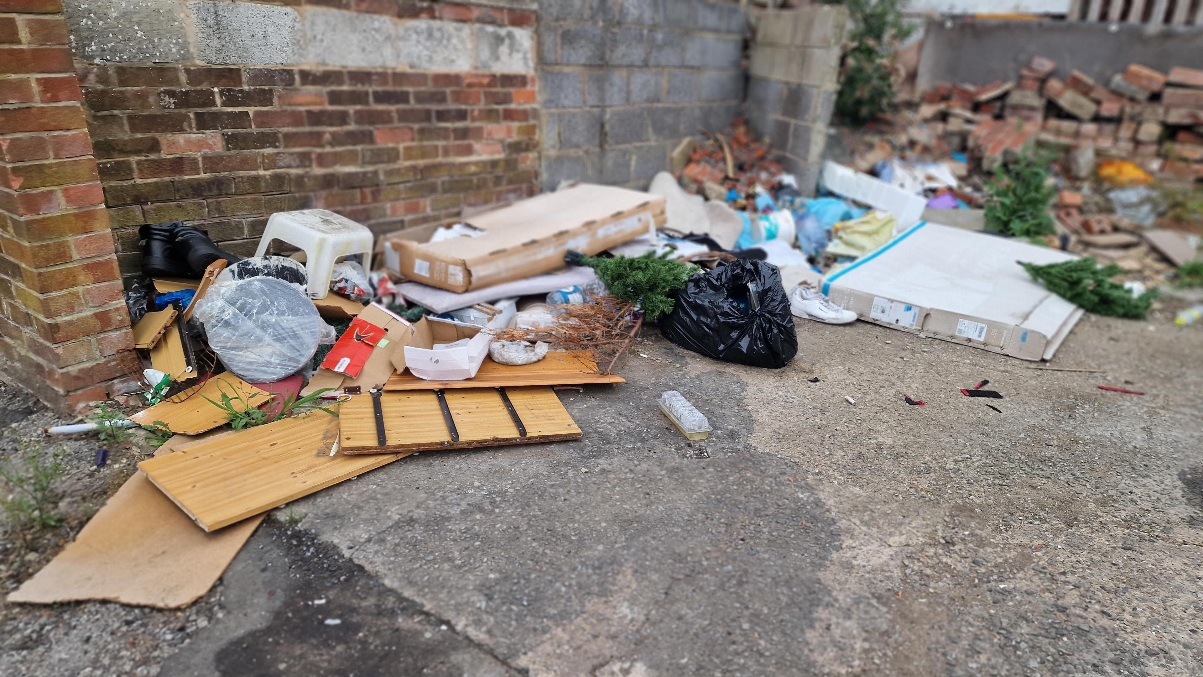 Household waste including wood, plastic and other items discarded on a concrete floor in front of a brick wall.
