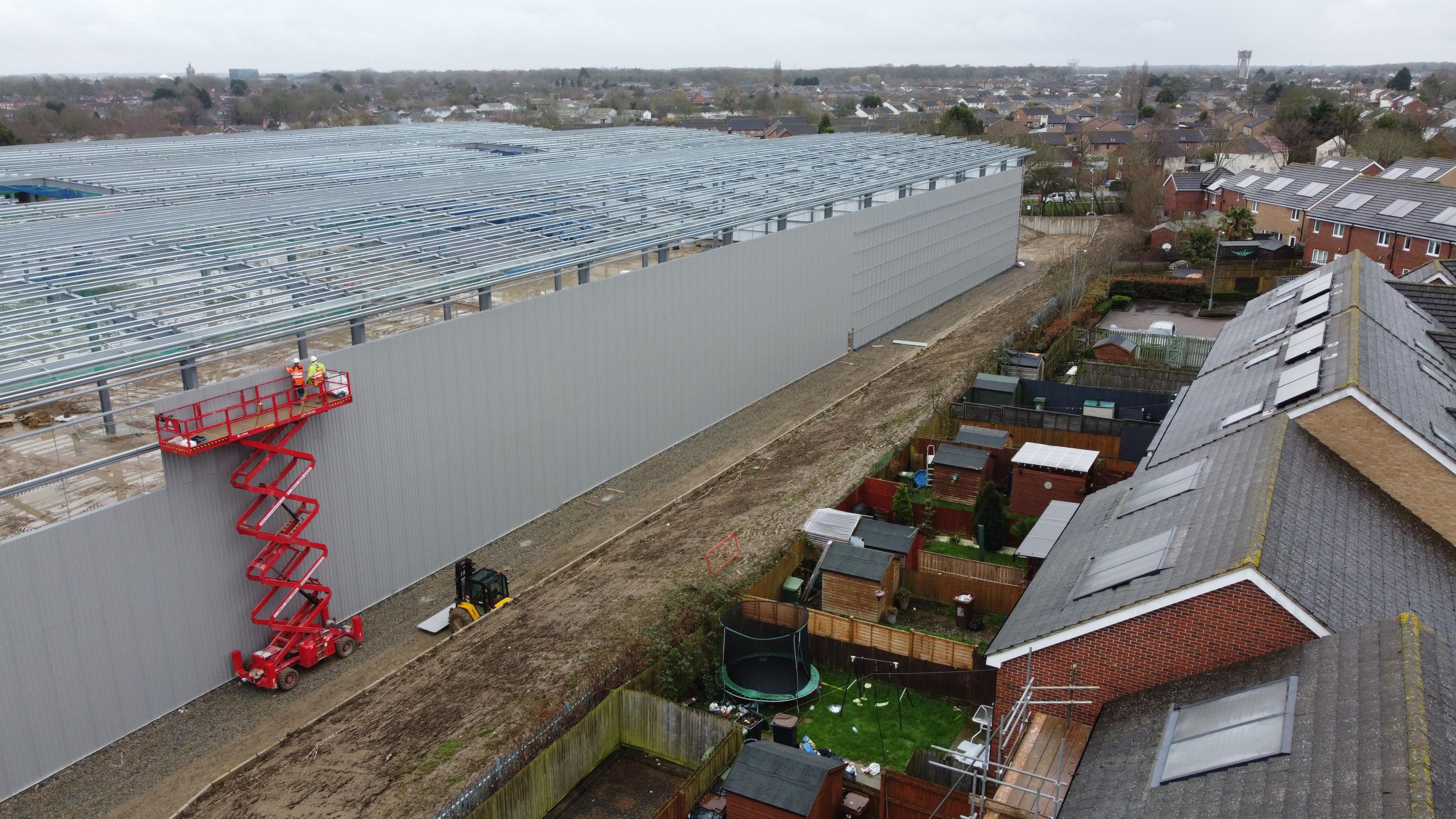 A drone shot of the incomplete warehouse - a grey structure that stretches across the frame several metres in front of a housing estate. The estate backs directly onto the warehouse land. 