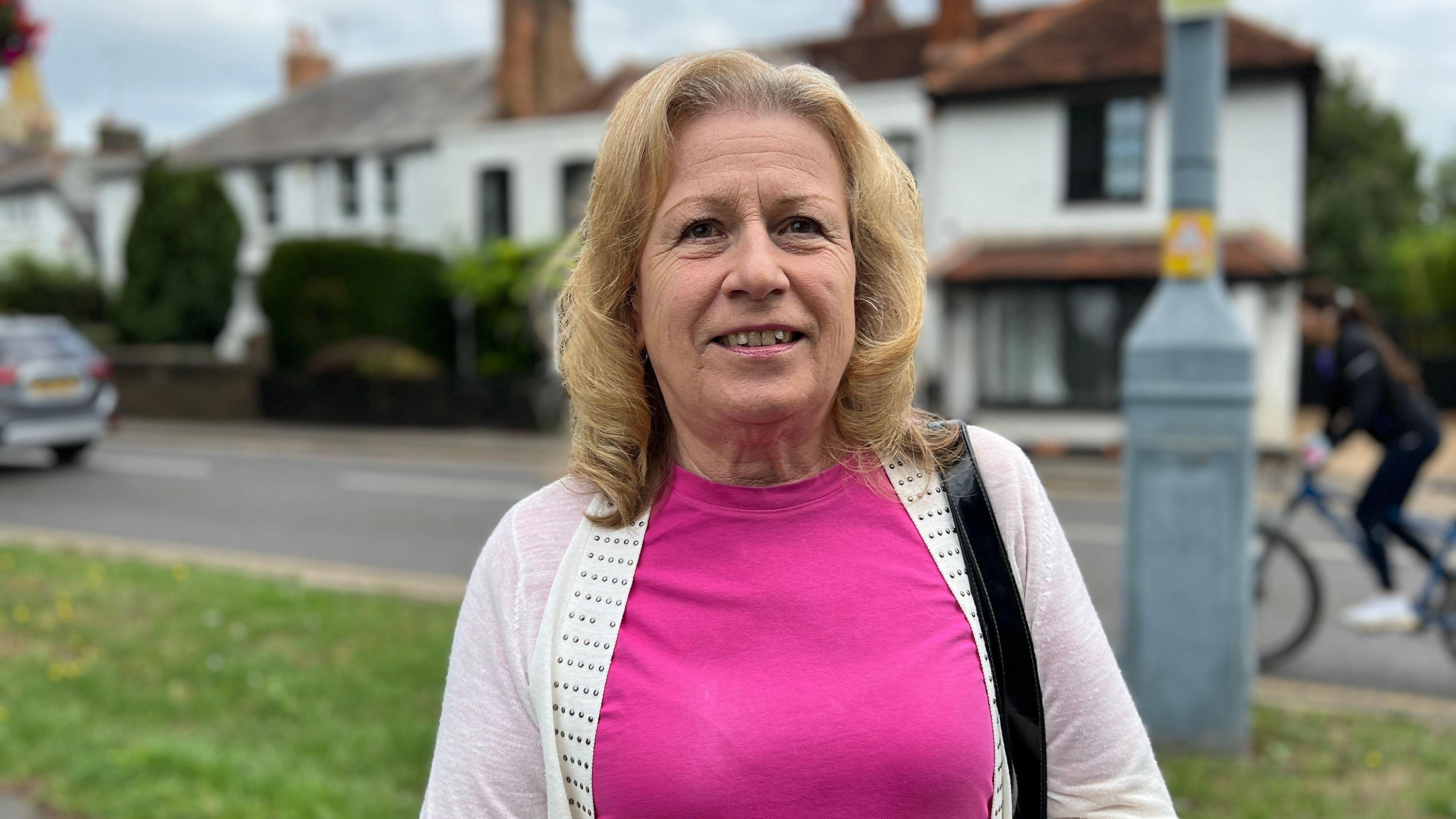 Zena Coccaro has shoulder-length blonde hair and is wearing a pink top and a white cardigan. There's a row of houses, a car, a cyclist and a road behind her. She's standing on a grass verge, and is looking at the camera and smiling.