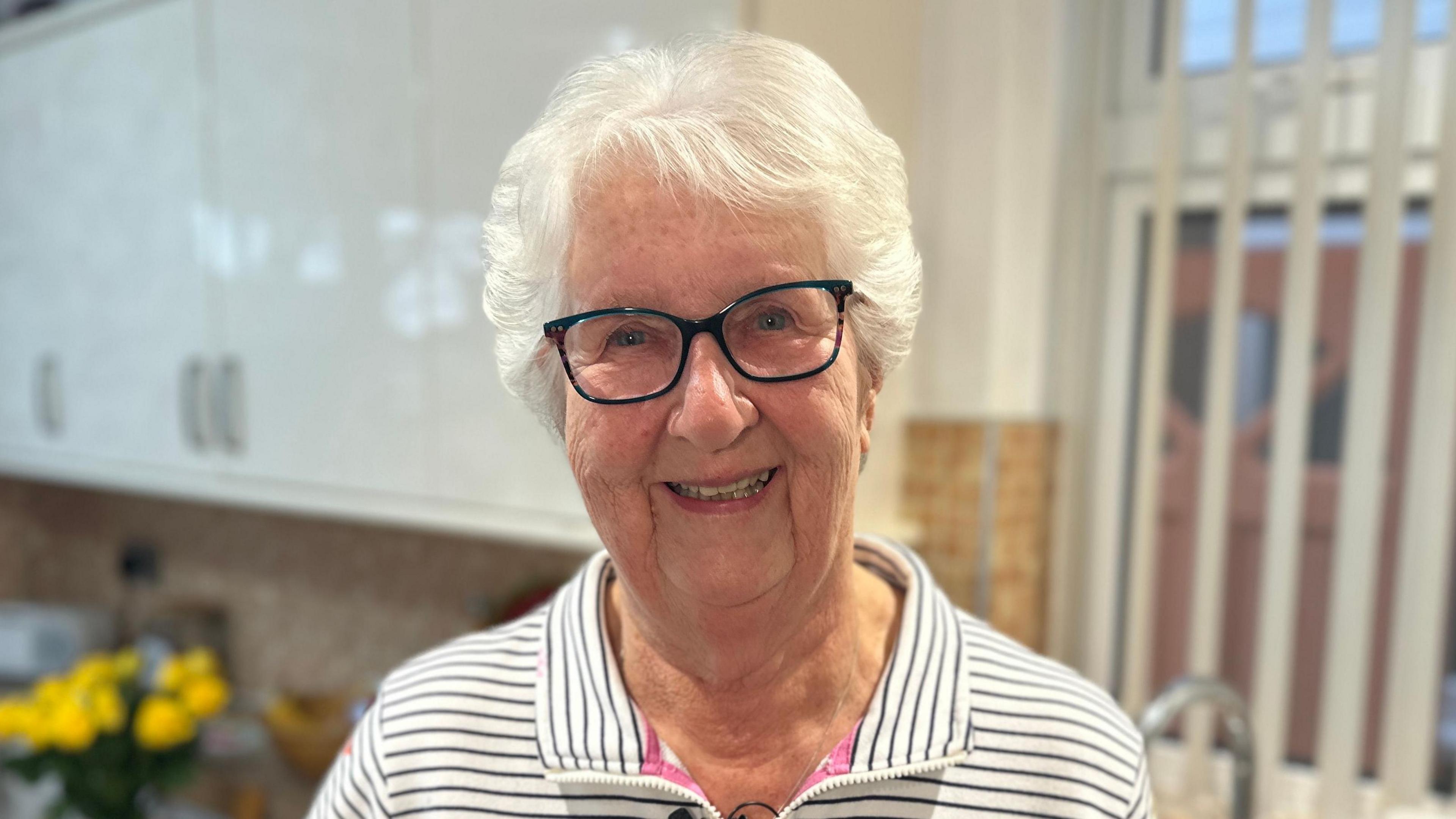 A grey-haired lady wearing glasses smiles at the camera inside a kitchen. She is wearing a stripy top.