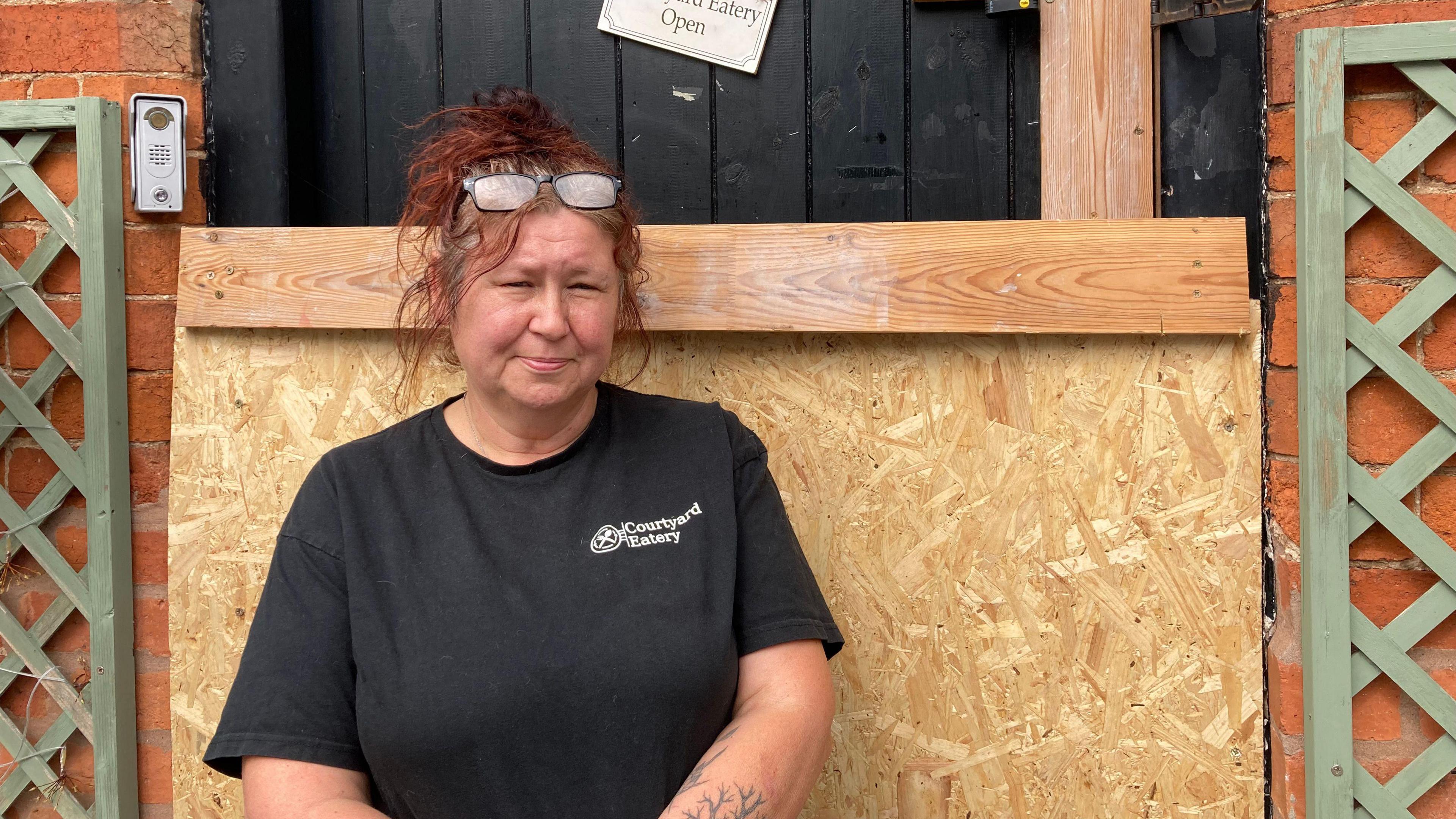 Woman with glasses on top of her head and she is wearing a black t-shirt with the words 'courtyard eatery' printed on the right hand side. She is standing in front of a boarded-up door.