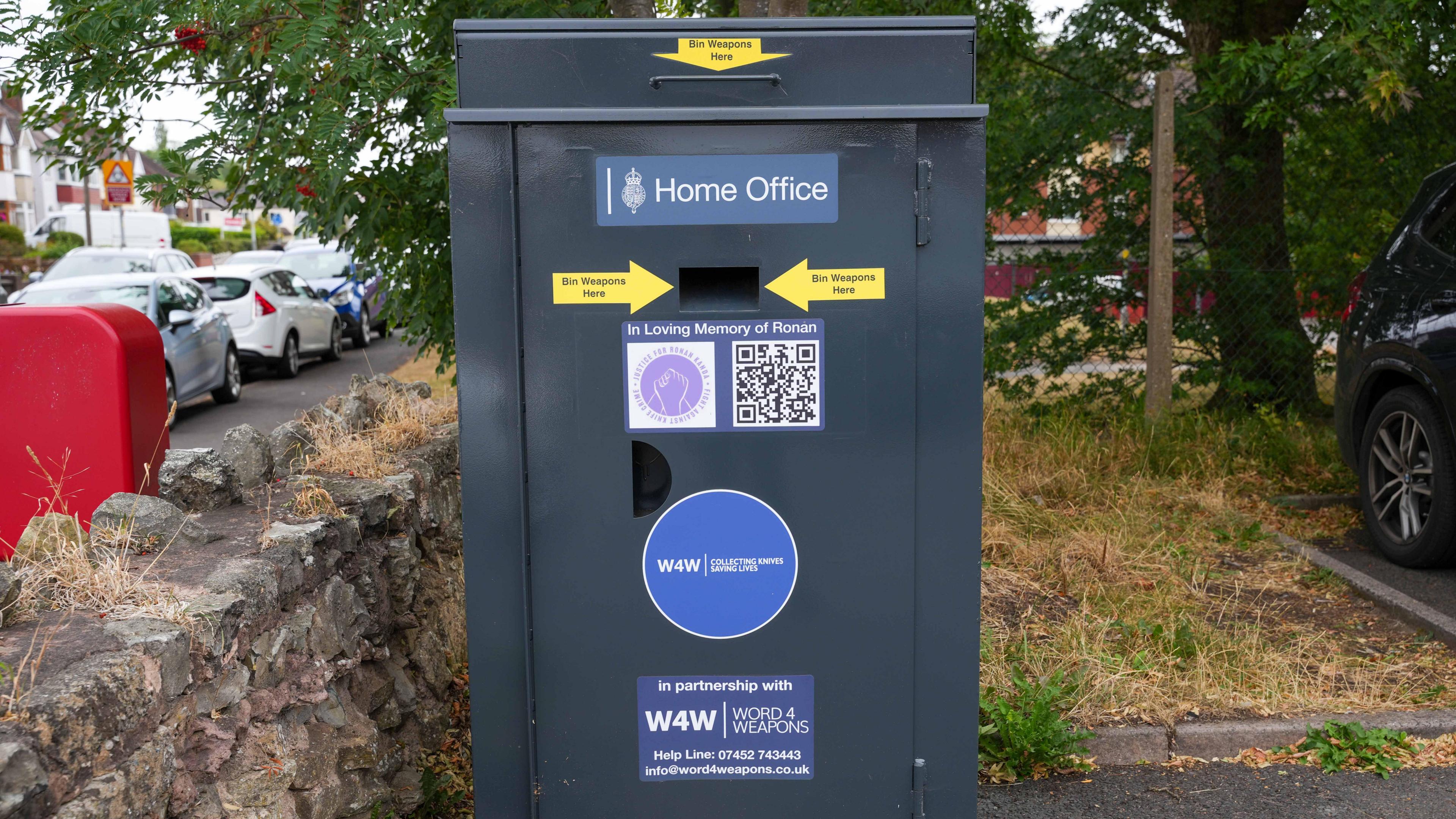 A large grey weapons bin which has a slot on it for people to abandon their weapons in. There is a large Home Office sticker on it and another sticker saying 'In Loving Memory of Ronan' with a QR code on it.