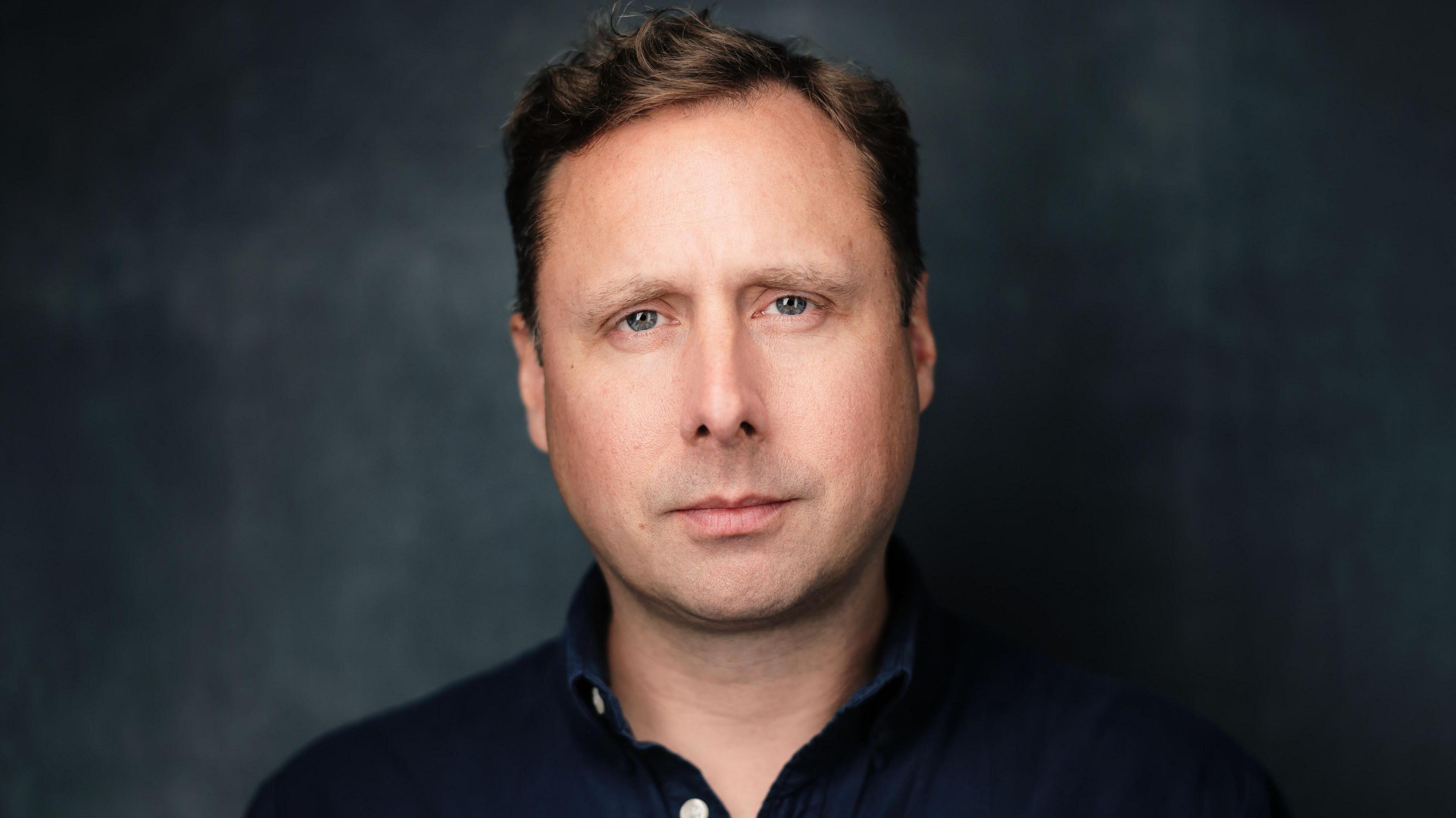 A headshot of Adam Graham, who is wearing a navy shirt. He has short brown hair and is looking at the camera without much expression. Behind him is a blurry grey and black background.