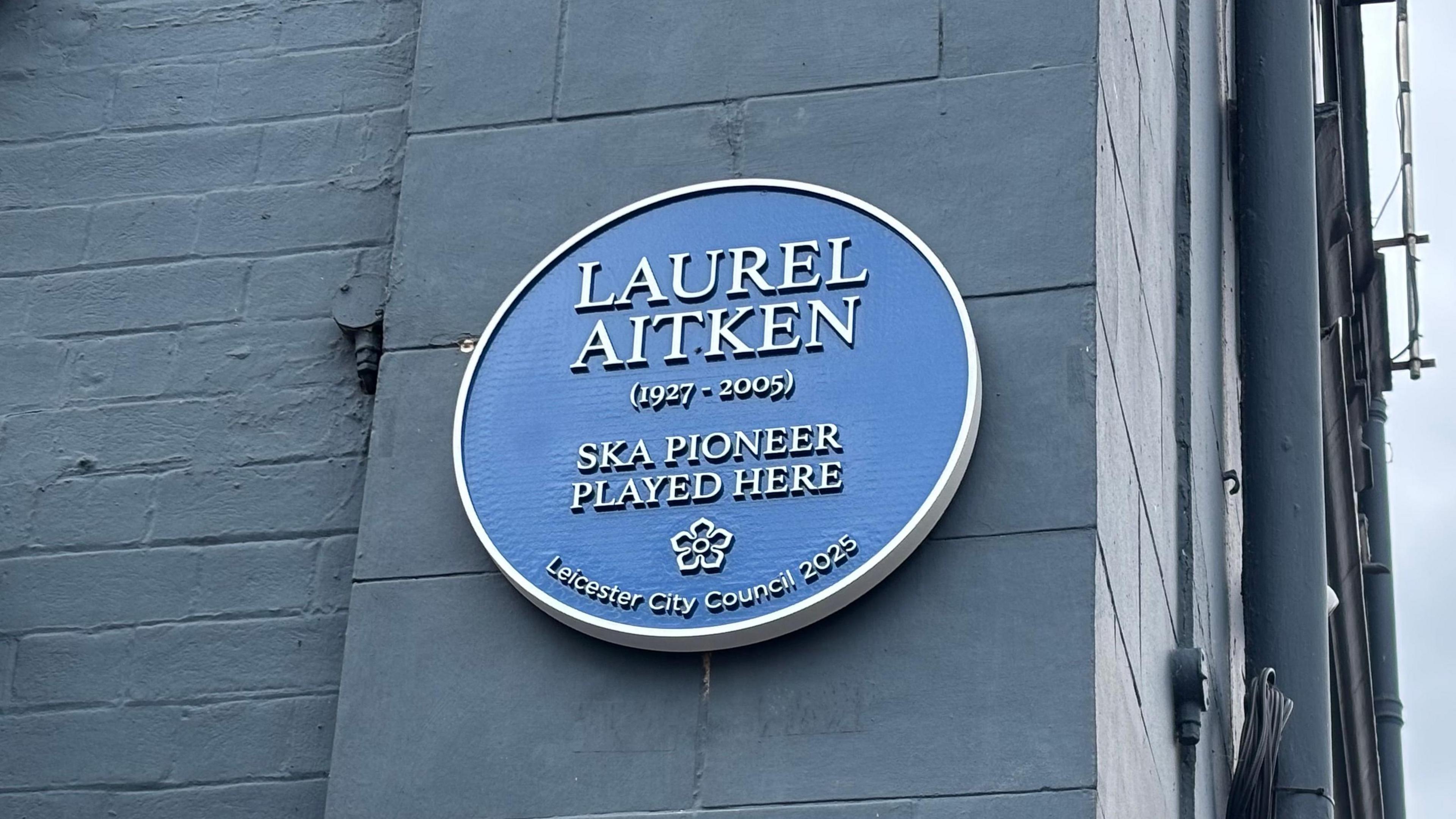 A blue plaque with Laurel Aitken on a grey-painted brick wall.