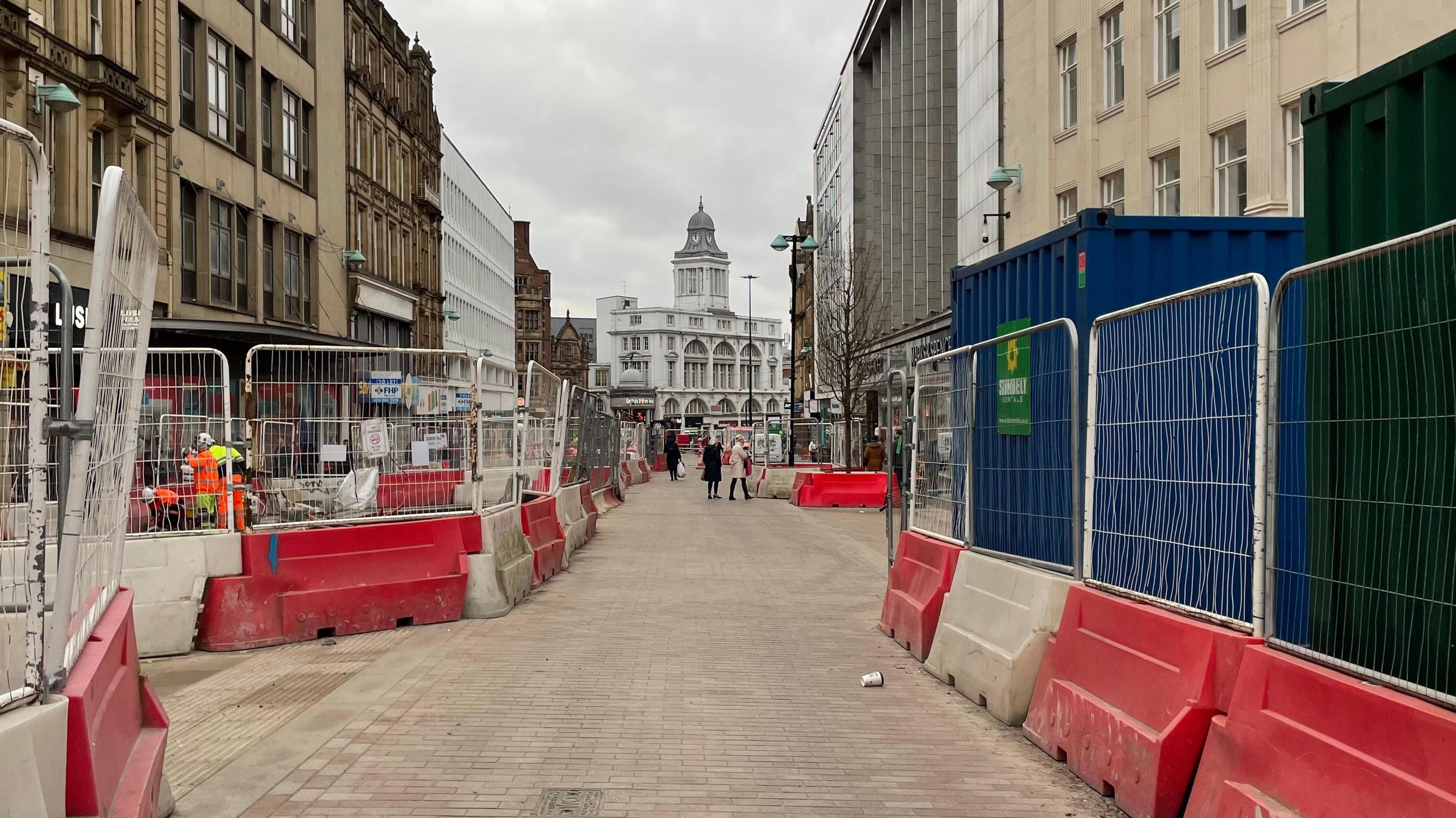 A pedestrianised street with red and white plastic barriers and fences. Large office buildings on both sides.