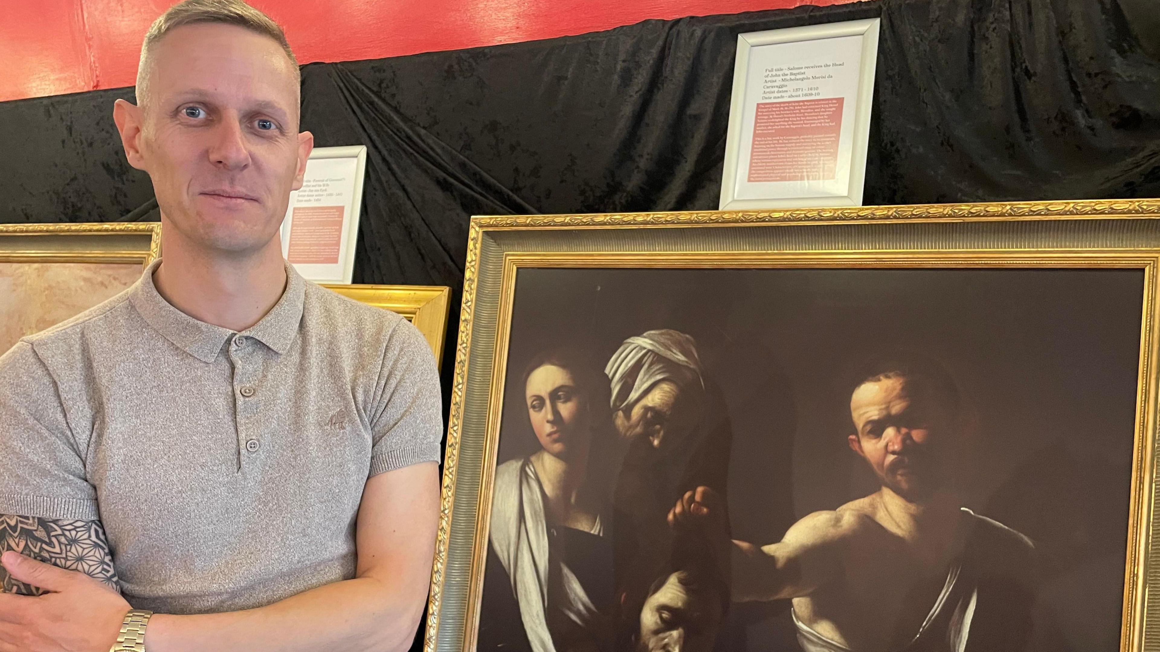 A man stands facing the camera beside a Caravggio painting. He is smiling and wearing a grey polo top