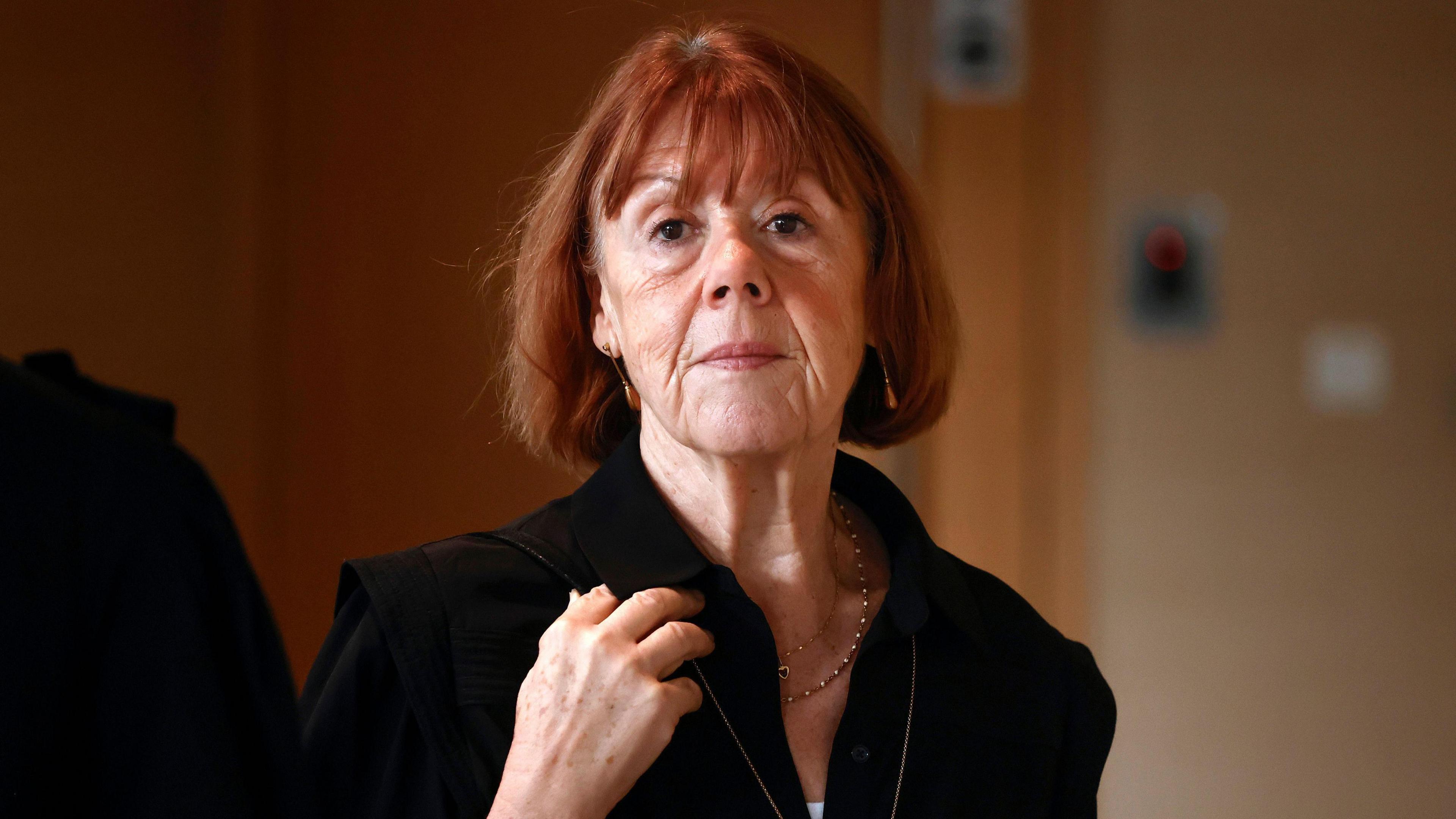 Gisèle Pelicot, a woman with red hair in a black top, looks at the camera. She is not smiling