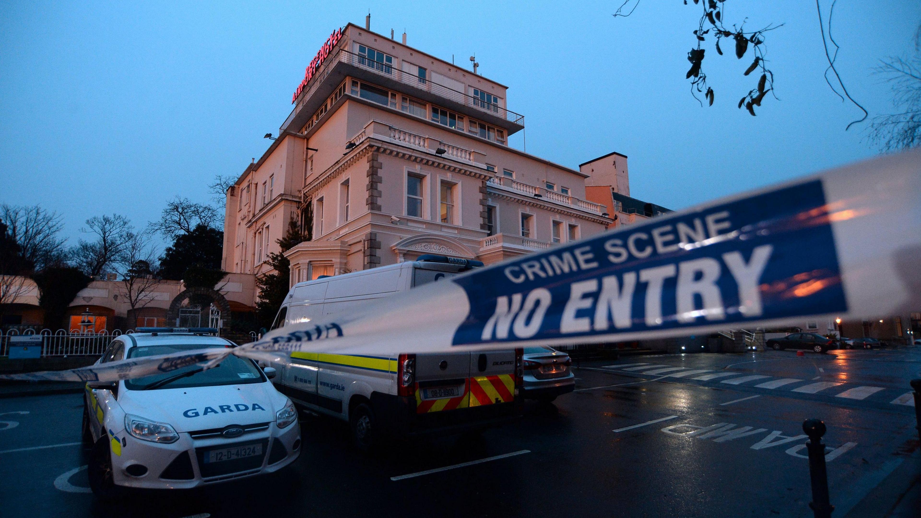 A garda (Irish police) cordon outside the Regency Hotel in Dublin in 2016.  A white and blue liveried garda van and car are parked outside a four-storey hotel at dusk on a wet evening.  A white a blue cordon tape stretches across an entrance to the hotel car park. 