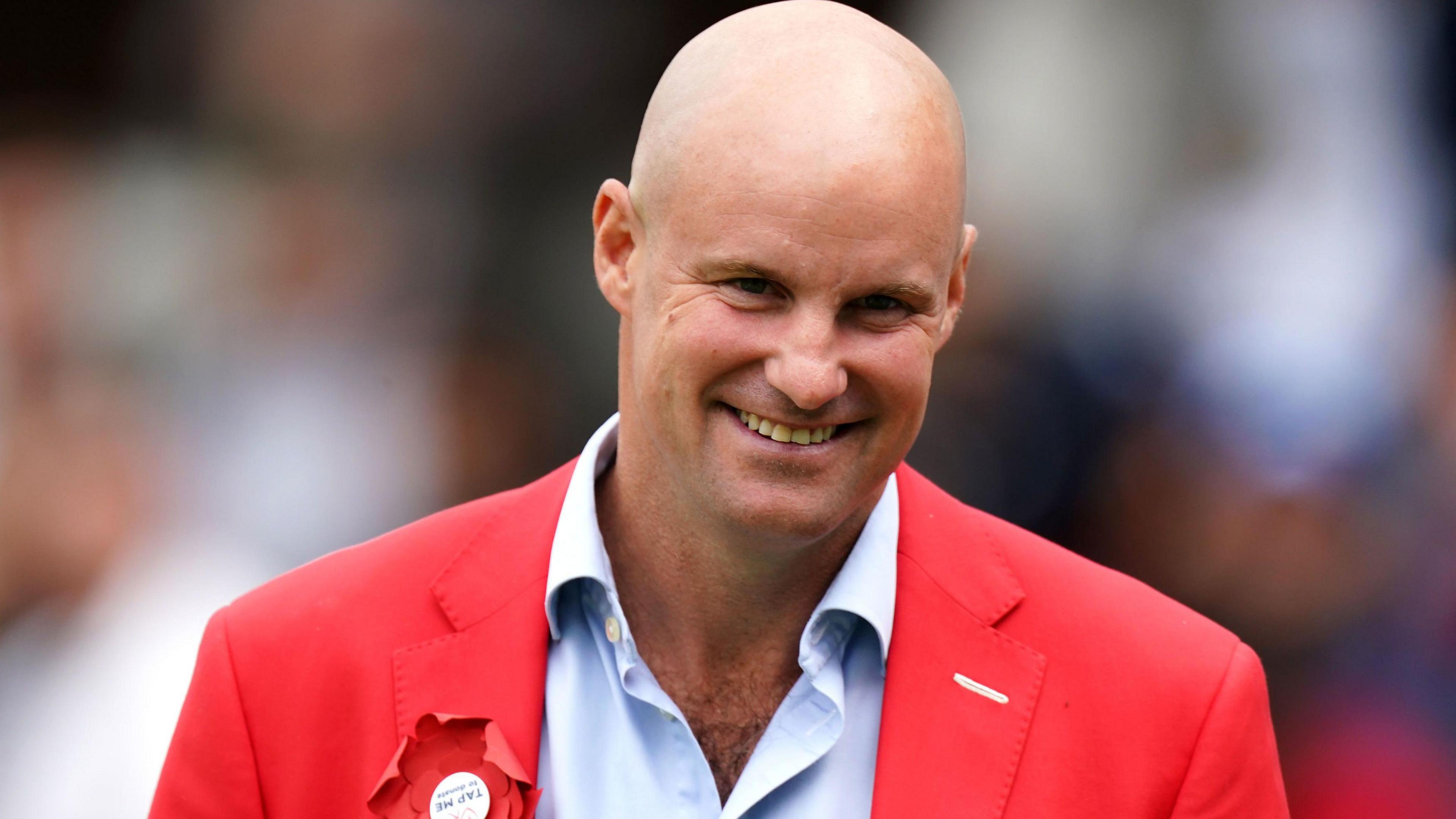 Andrew Strauss with a bald head smiling at the camera. He is wearing a pale blue shirt with a red blazer that has a red rosette on the left.