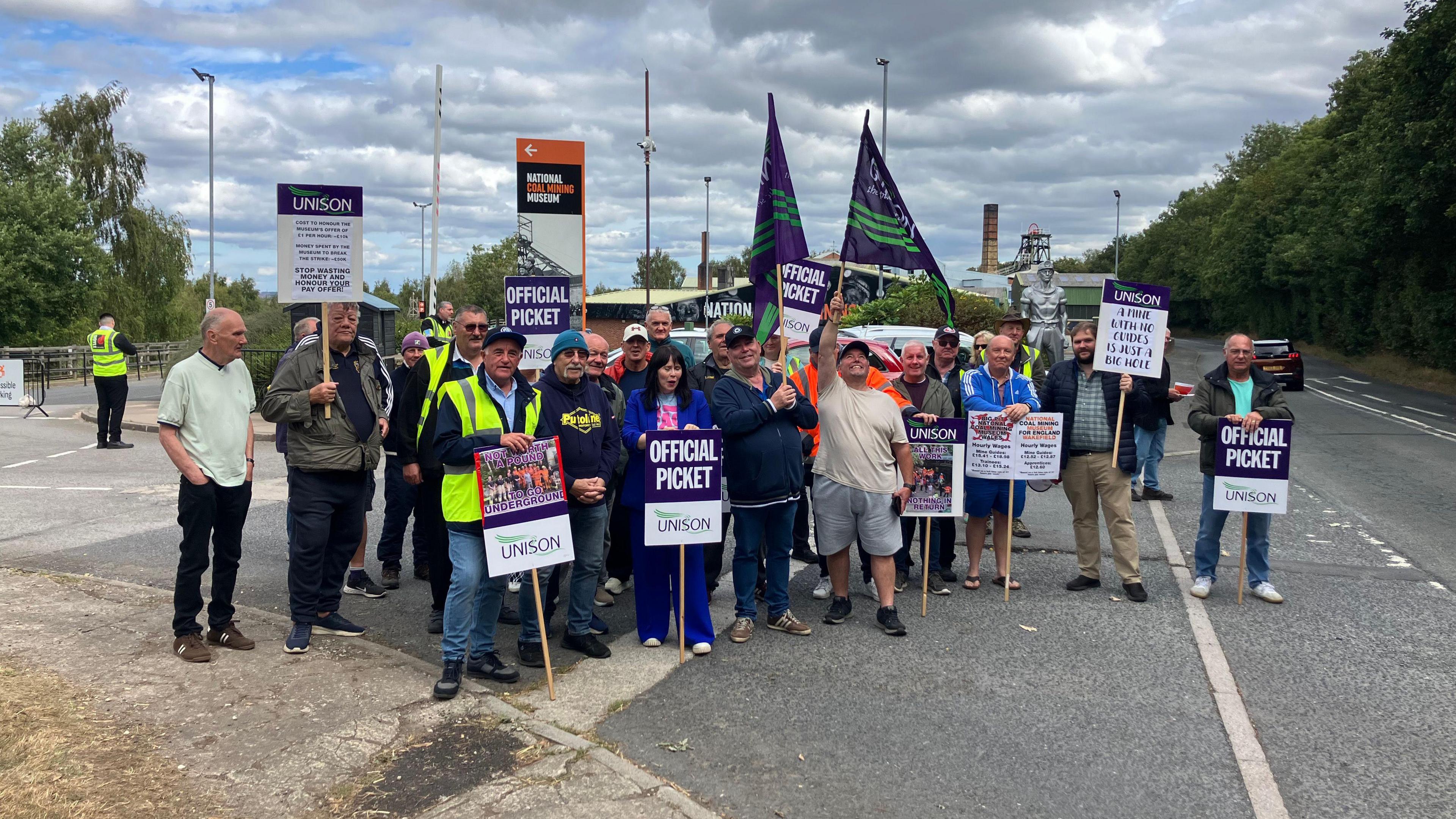 A group of workers stand in front of the National Coal Mining Museum. They wave banners and plaquards and wear high visibility jackets.