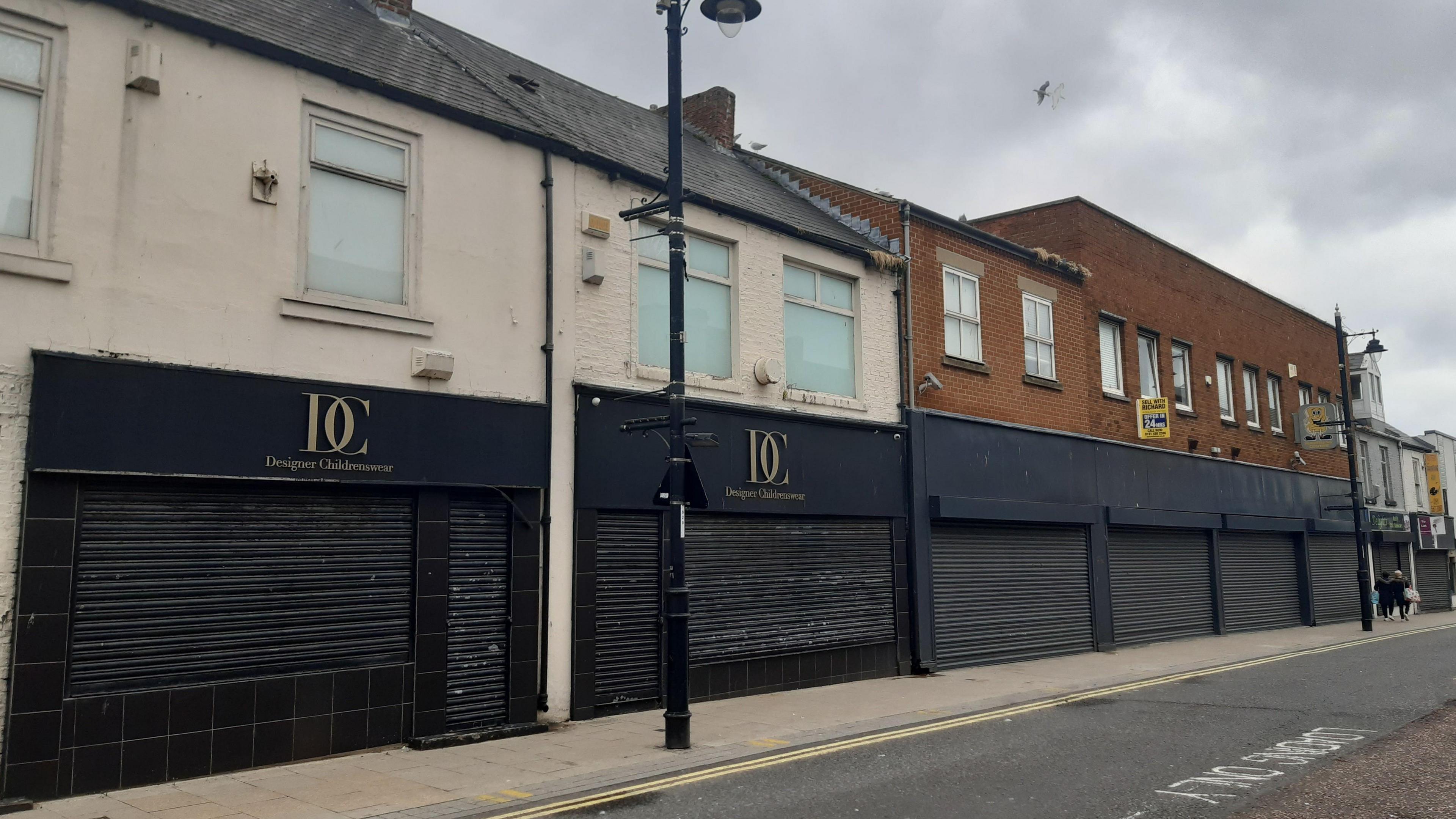 Street with former retail units, the first two on the right have white upper storeys, while the one of the left is brick. All have closed black shutters covering both the downstairs windows and doorways.