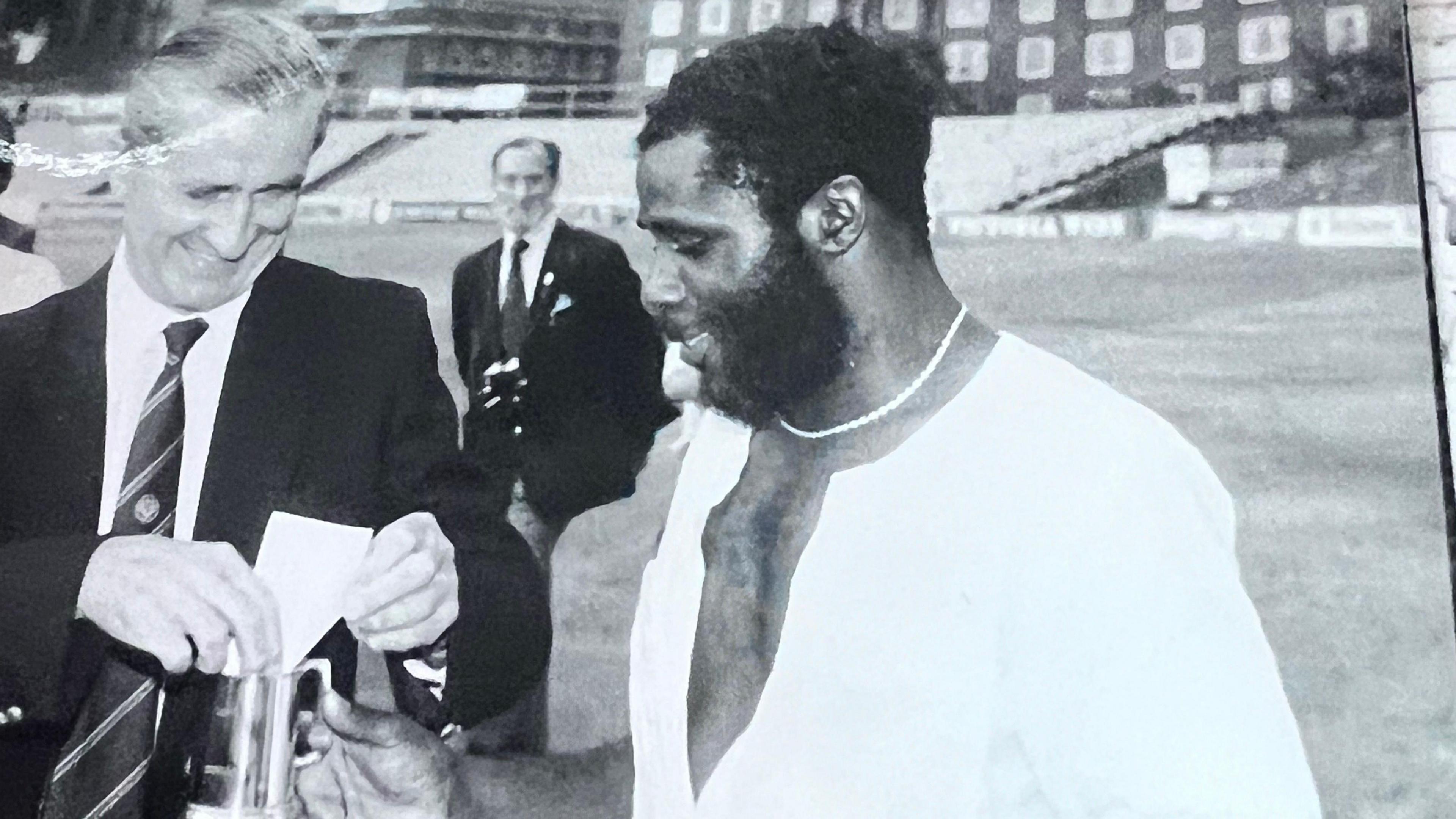A black and white image from 1982, showing a West Indies Cricket player holding a silver cup, as another man places a piece of paper in it.
