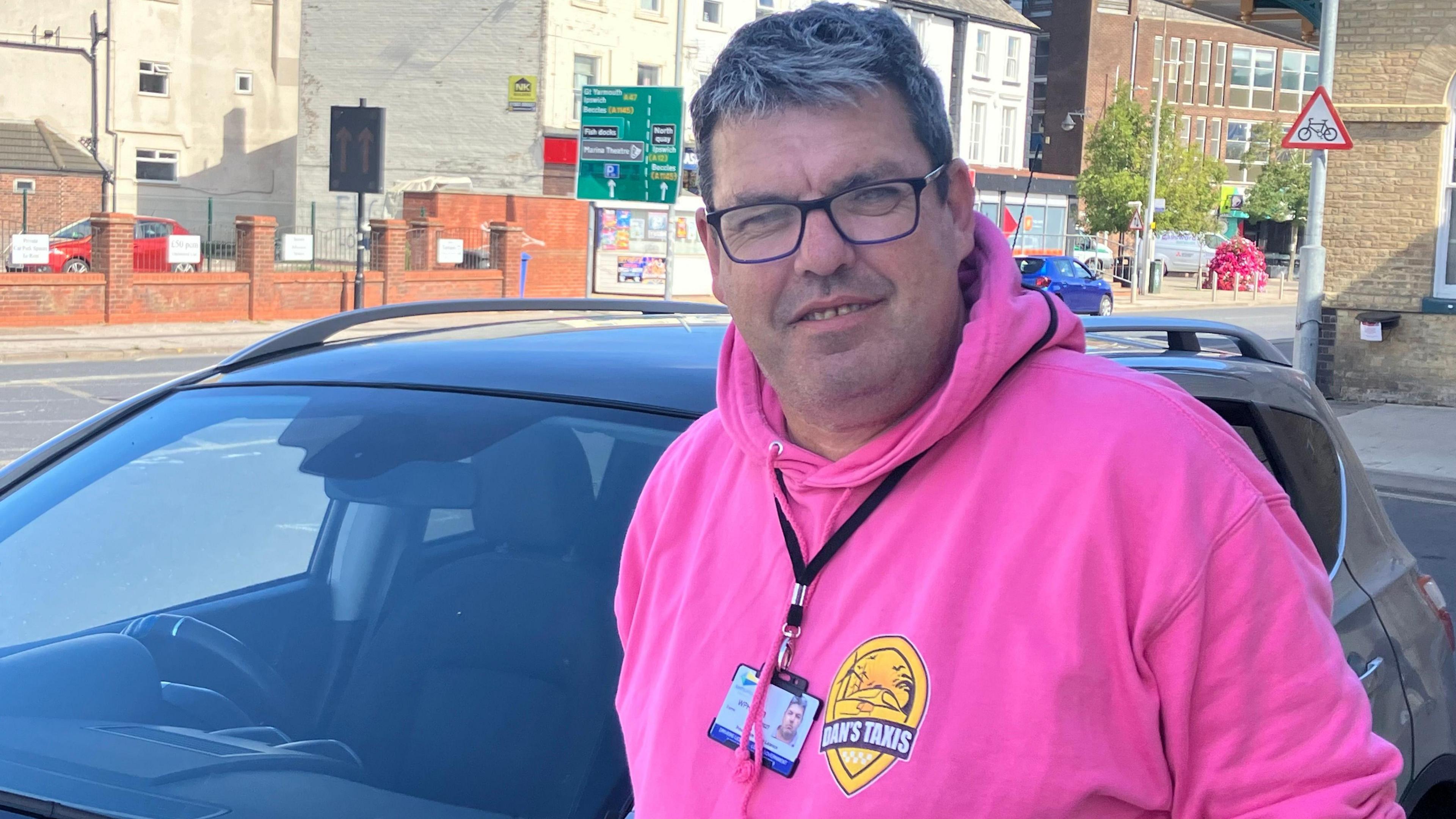 A taxi driver stands in front of his vehicle wearing a bright pink top, glasses and an identity pass around his neck