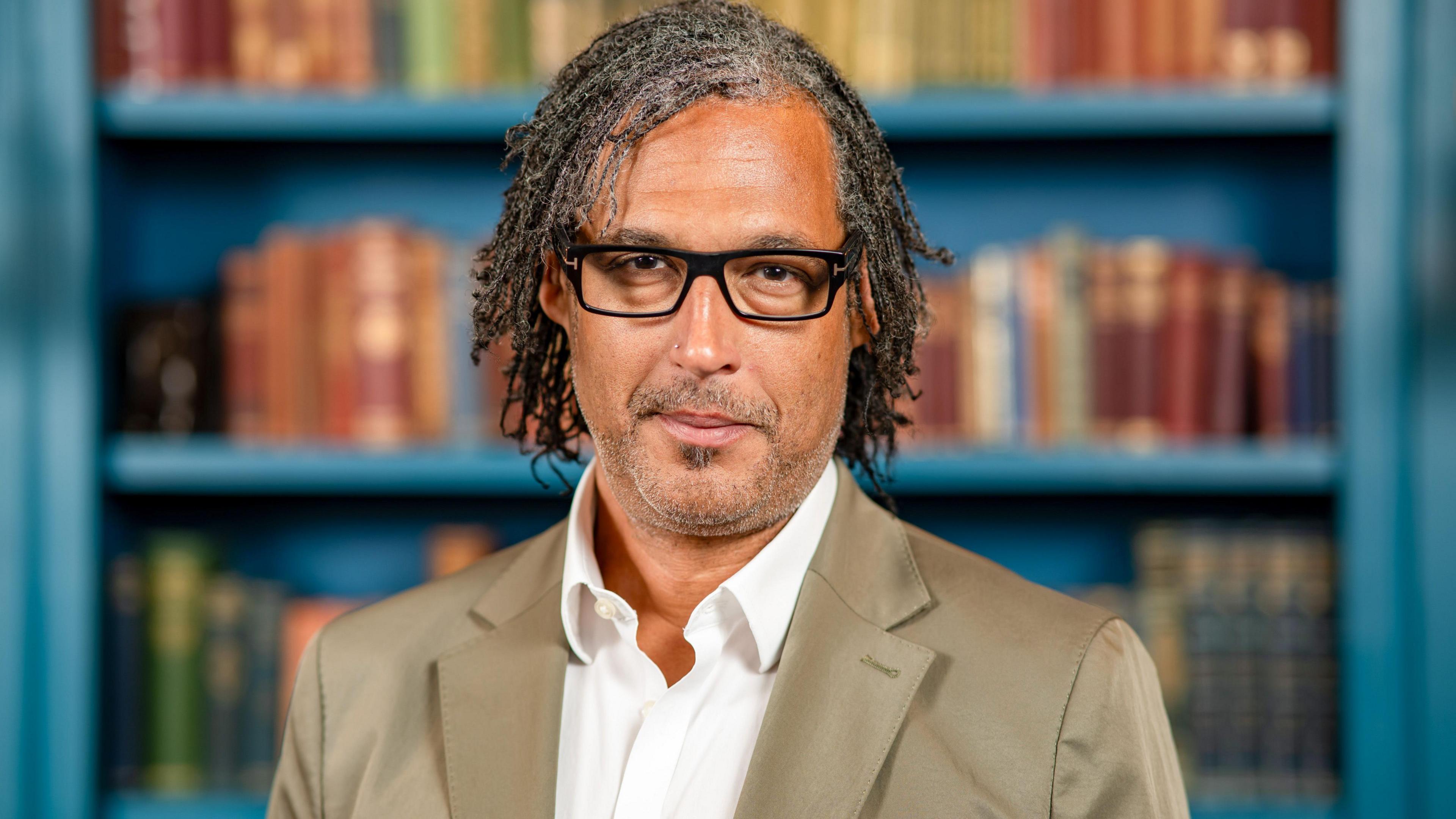 David Olusoga. He has dark hair, stubble and glasses on. He is pictured in front of a blue book case, looking directly at the camera and smiling.