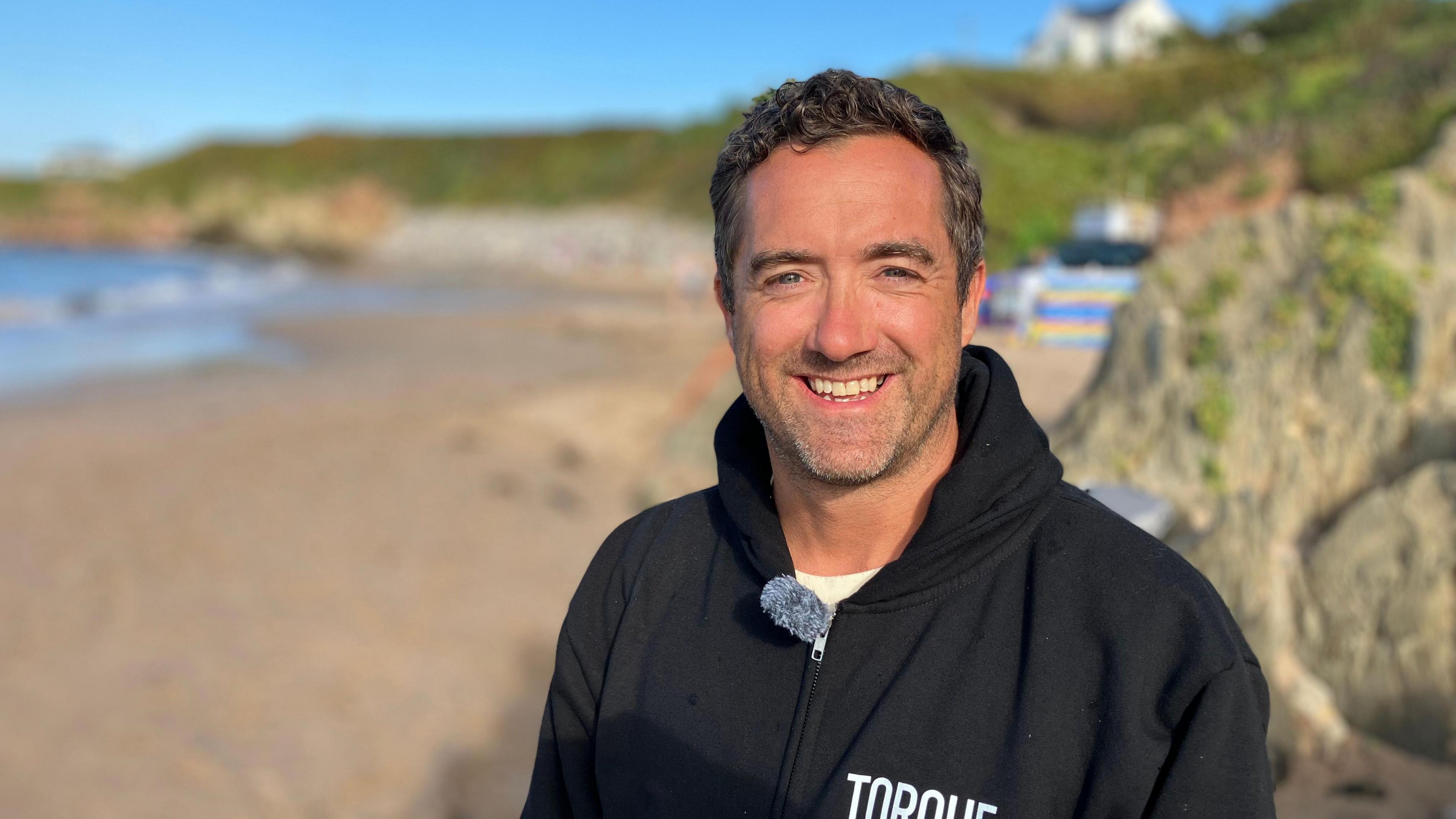 A man stands on a beach on a sunny morning, he is smiling and wearing a black fleece. You can see the beach, colourful windbreaks and the sea in the background.