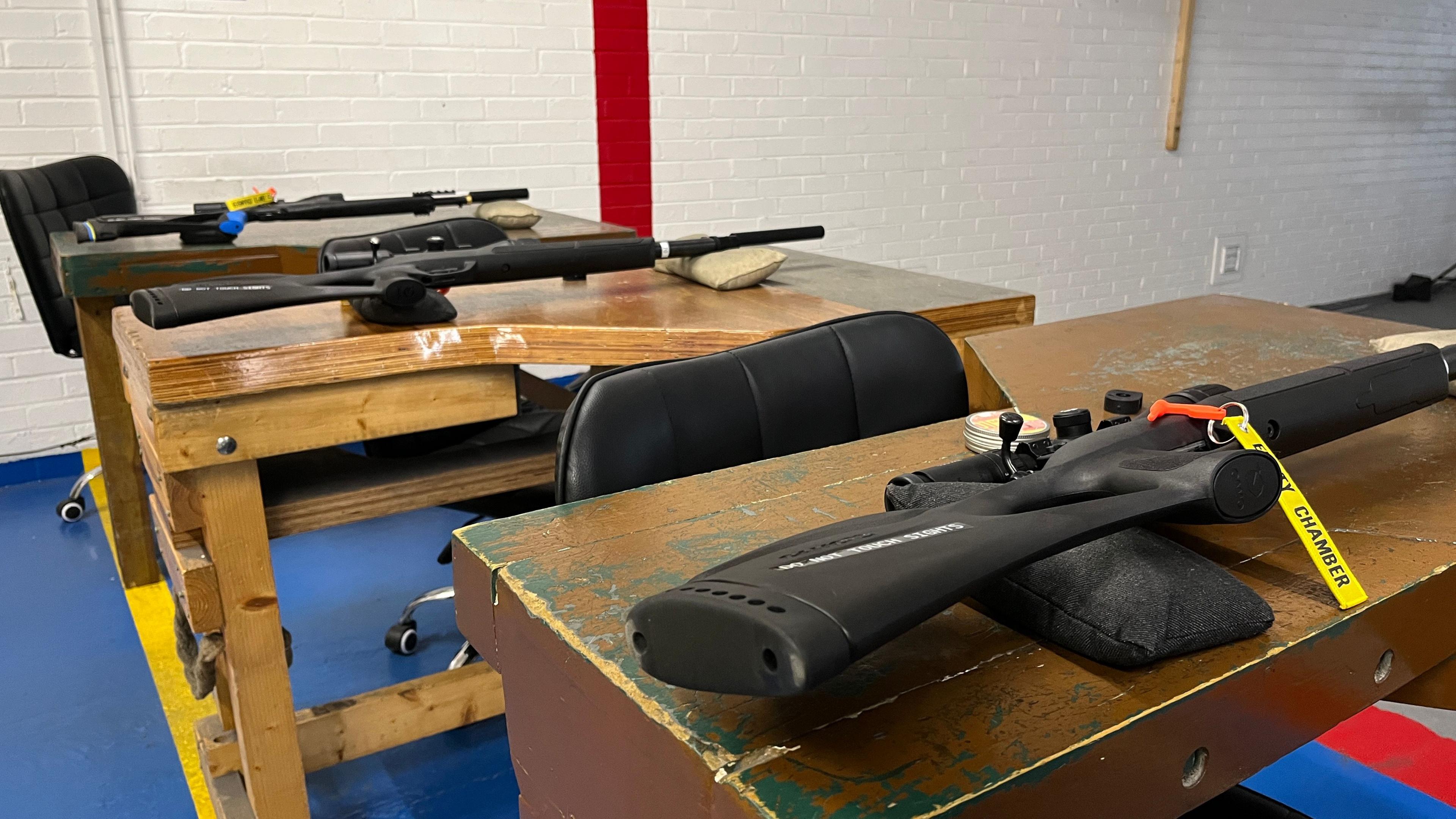 Three rifles have been lay on three wooden tables in a white bricked room.
